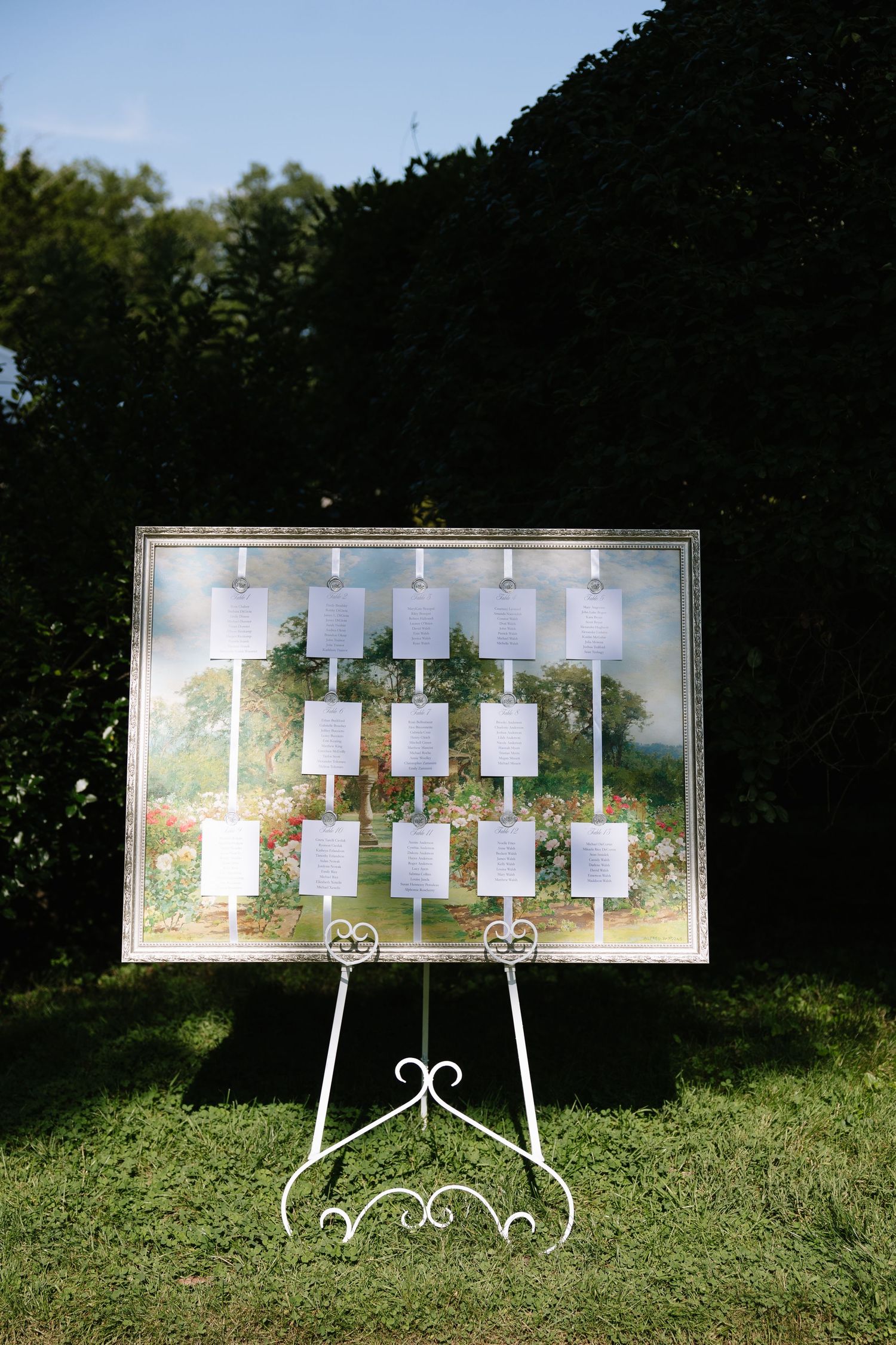 Elegant white framed wedding seating chart display on ornate easel outdoors on green grass.