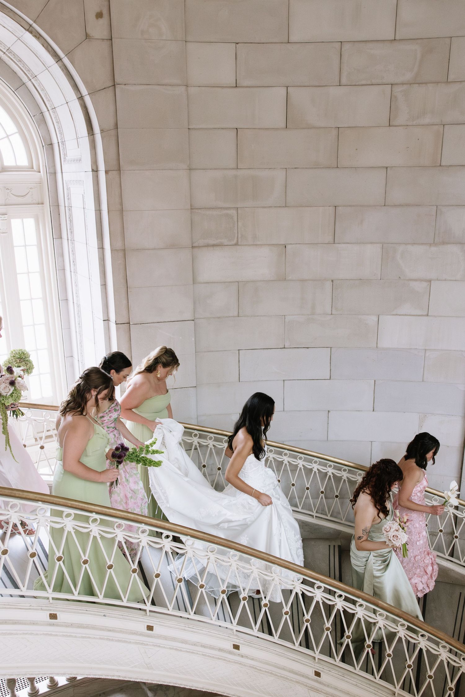 Bride in white gown walks up elegant staircase with bridesmaids in sage green dresses holding bouquets.