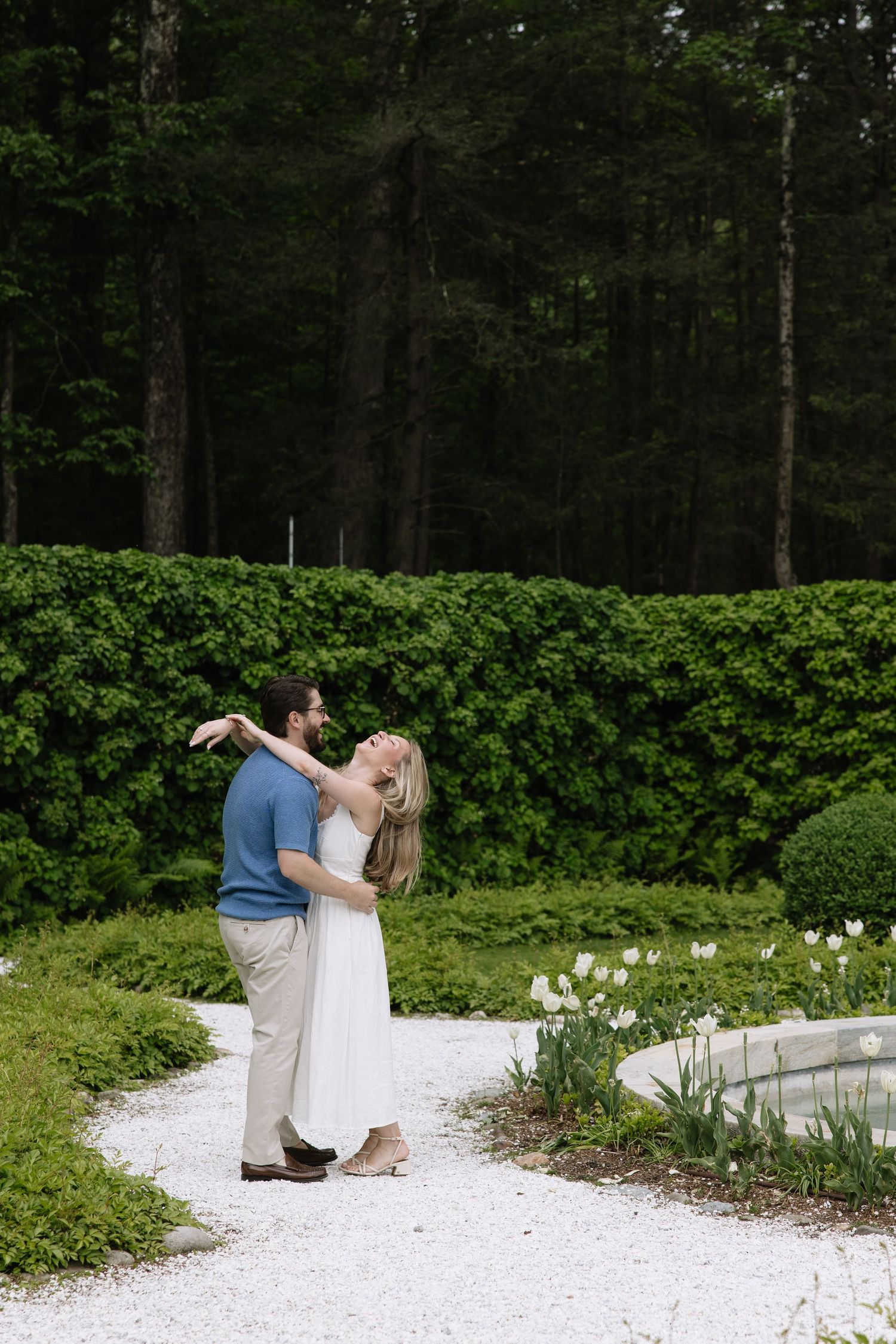 Couple dancing and embracing on a white gravel garden path surrounded by lush green hedges and white tulips.