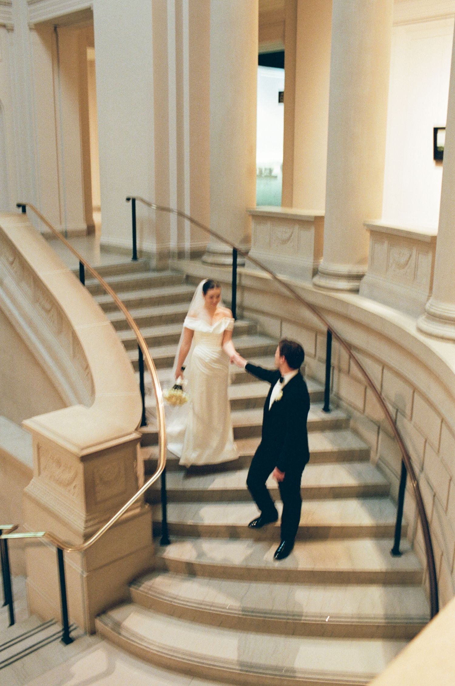 Bride and groom holding hands on elegant curved marble staircase in a grand wedding venue.