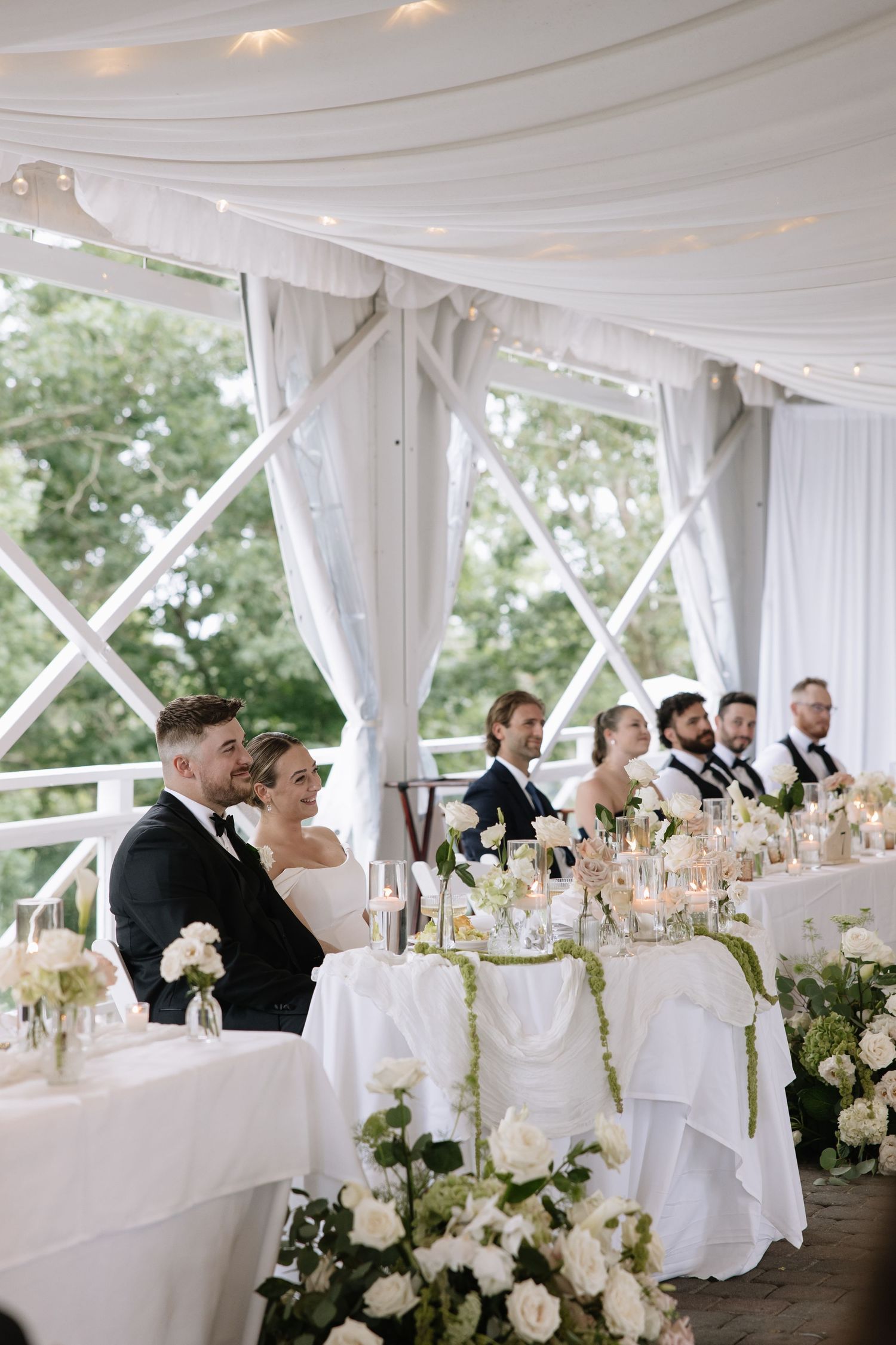 Bride and groom smiling at wedding reception under elegant white draped tent with white floral centerpieces.