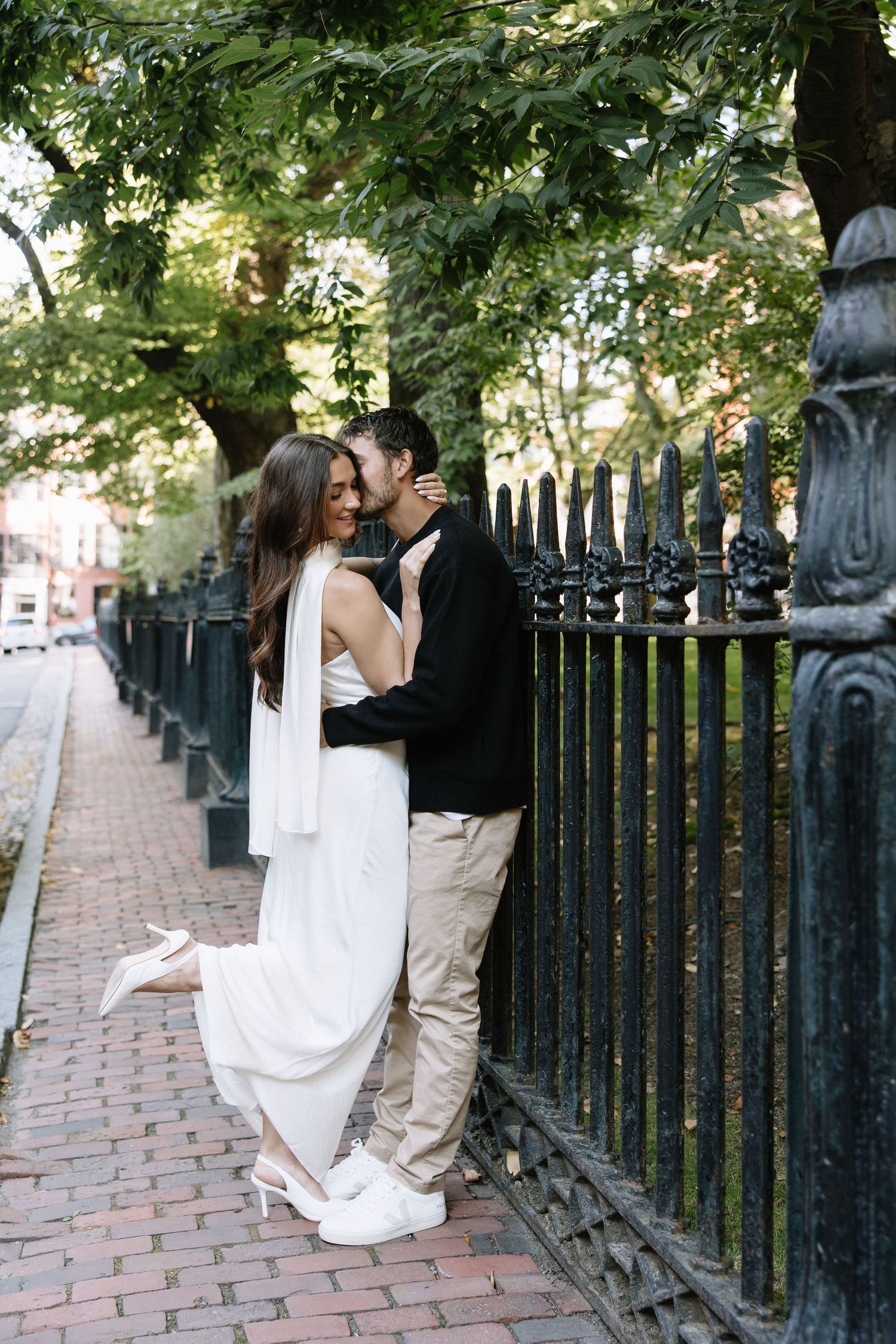 A romantic couple embraces against an ornate iron fence on a brick sidewalk in an urban setting.