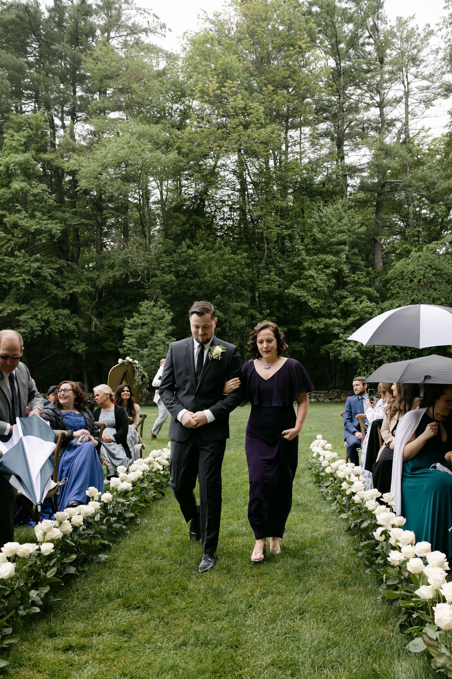 Wedding guests gather along a flower-lined aisle during an outdoor ceremony.
