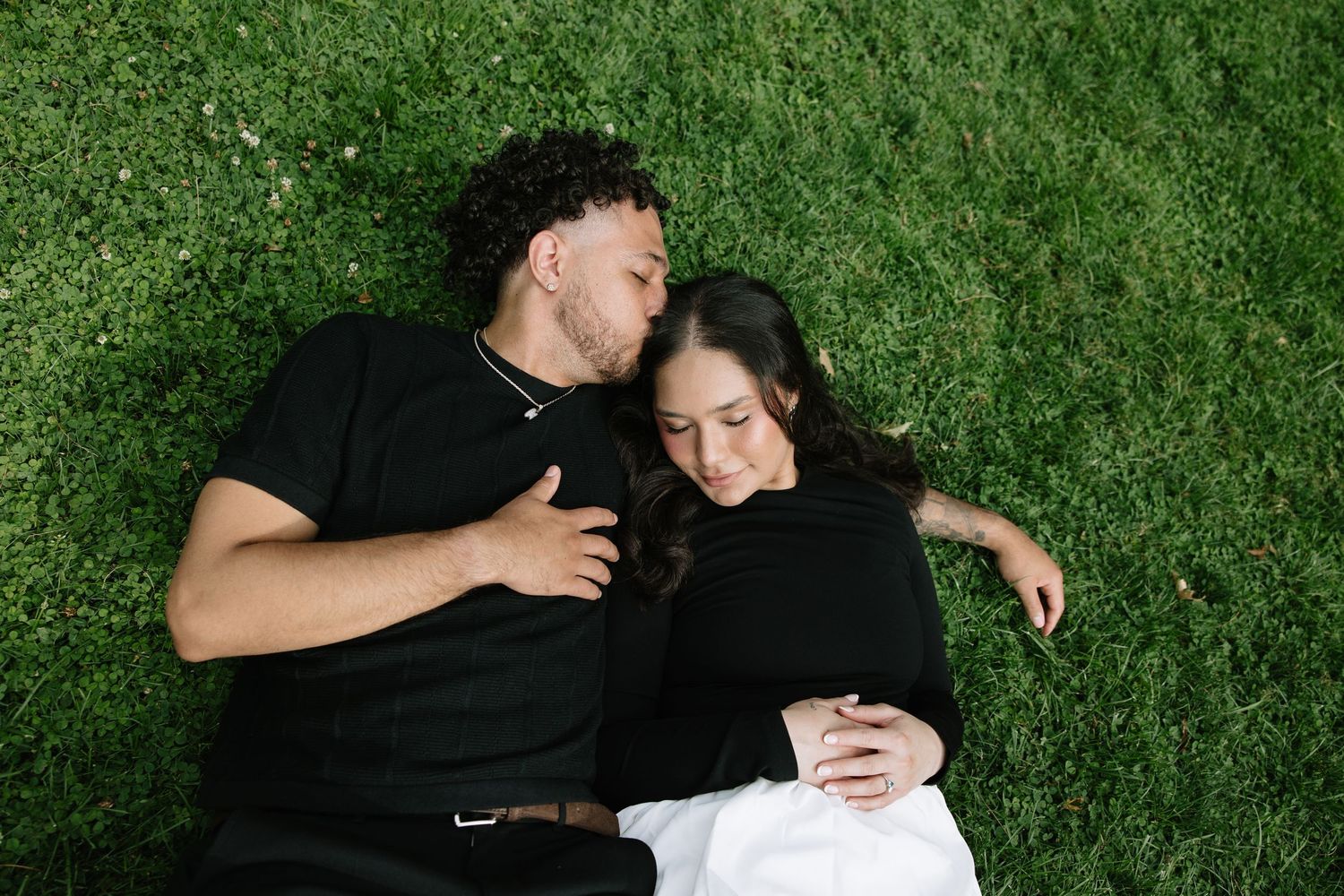 A couple in black clothing lying together on green grass in a romantic pose.