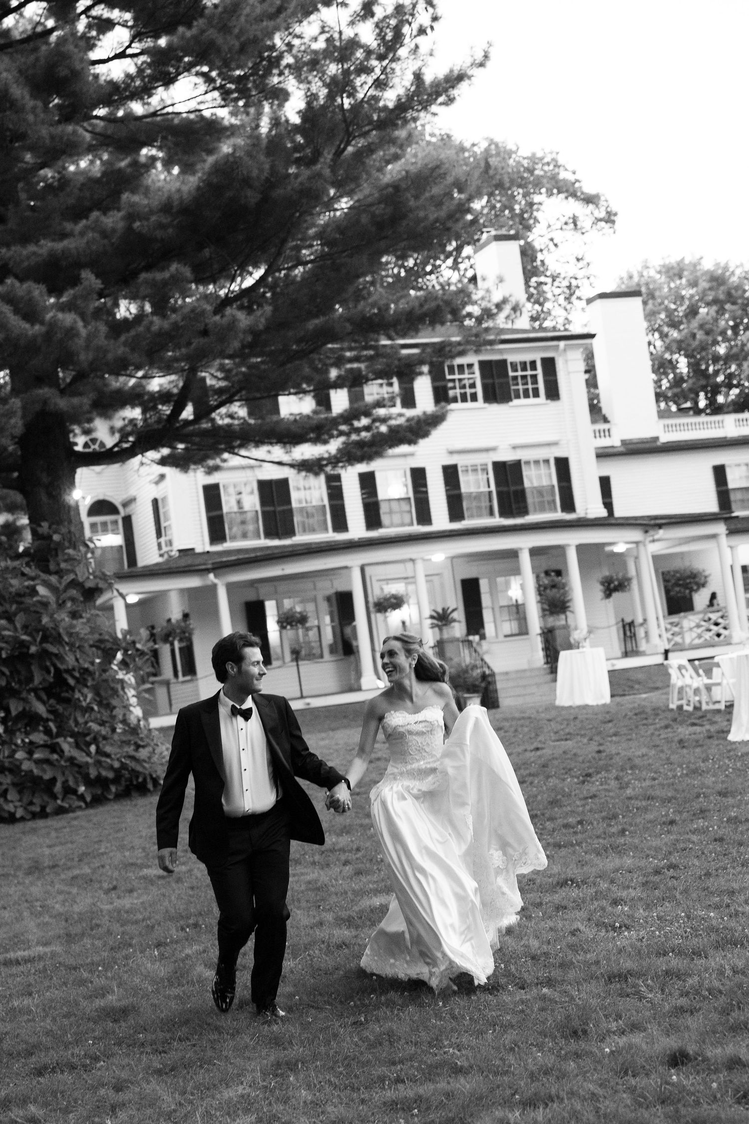 Wedding couple runs playfully across a lawn in front of a white colonial-style building.
