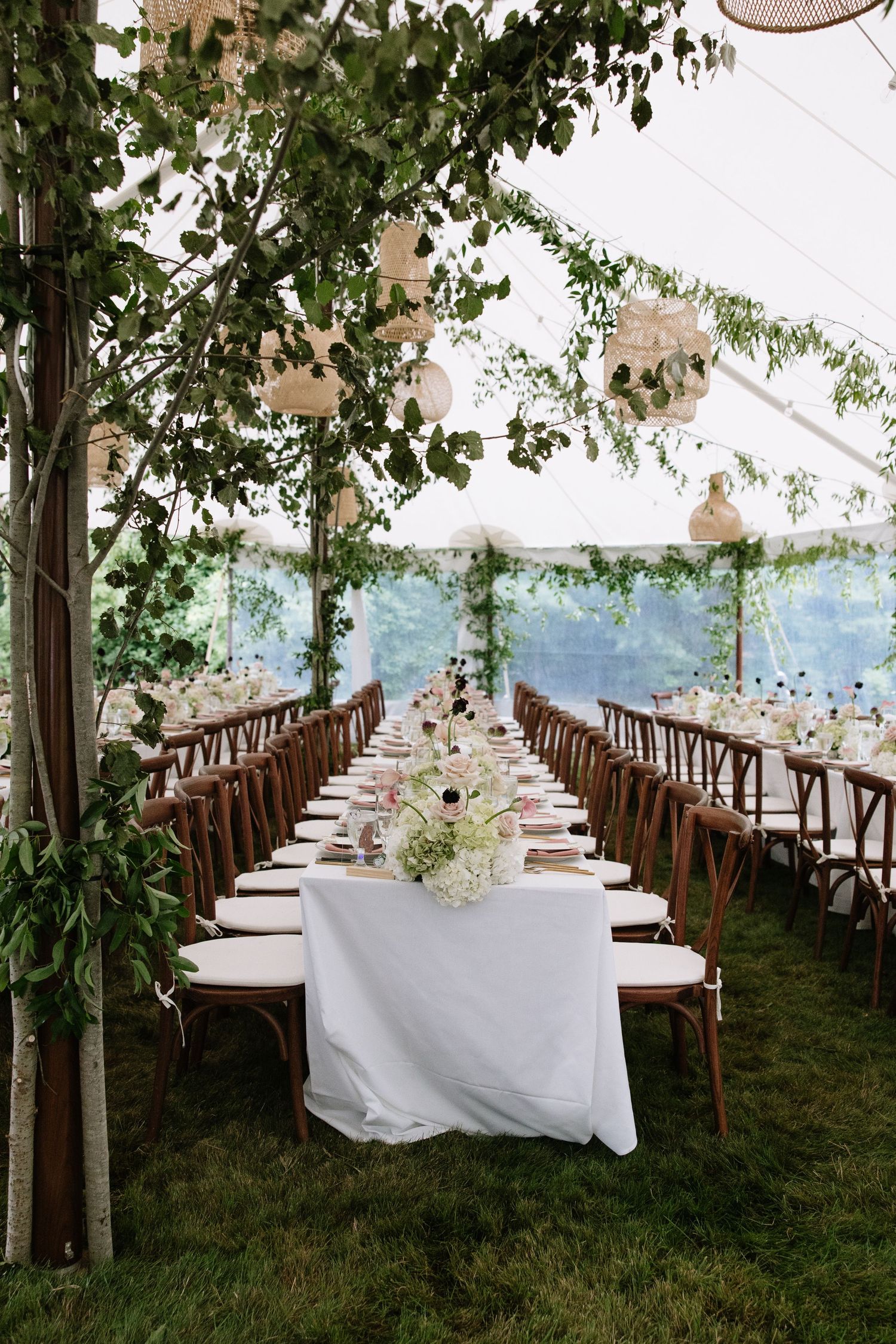 Elegant outdoor wedding reception under a white tent with long tables, white linens, floral centerpieces, and lush greenery.