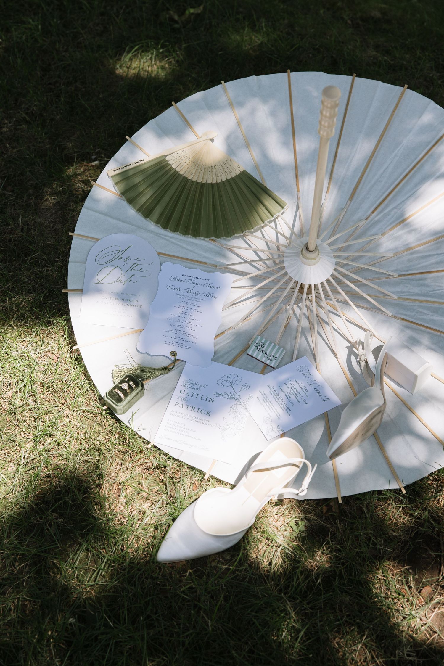 White parasol on grass with wedding stationery, green fan, and bridal shoes arranged elegantly for a wedding flat lay.