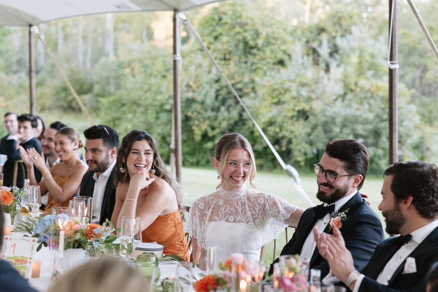 Bride and groom laughing at wedding reception head table under elegant tent with floral centerpieces.