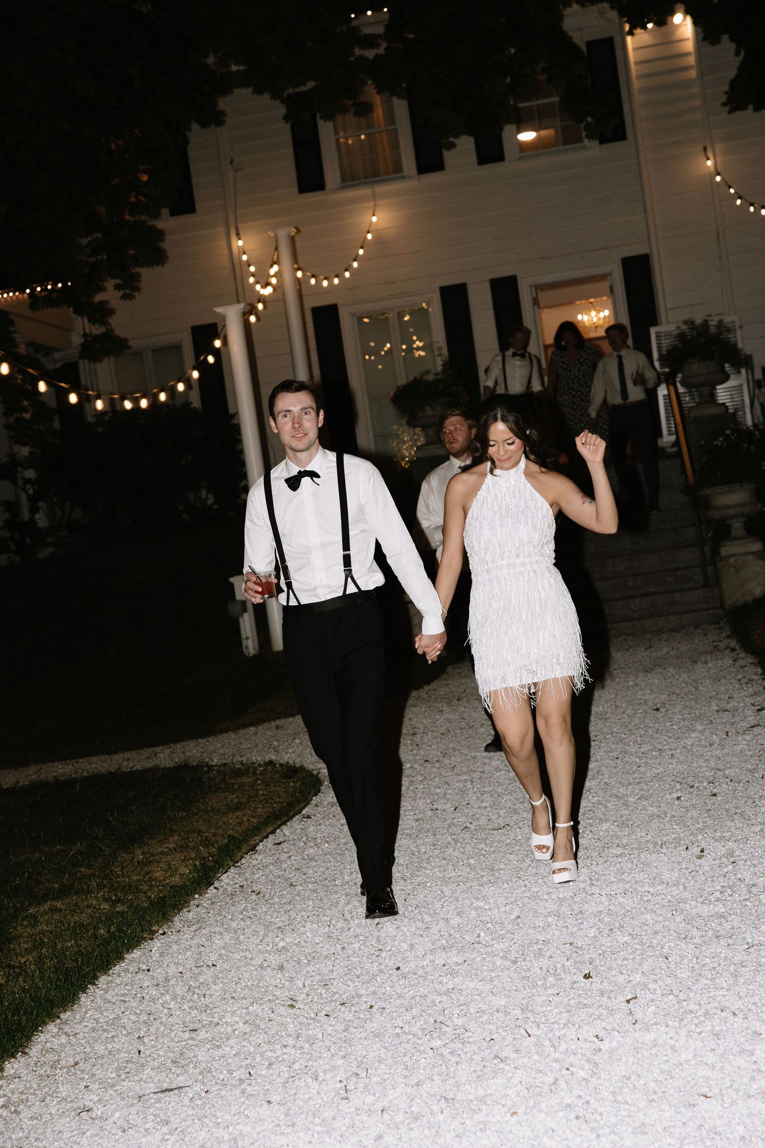 Couple in wedding attire dancing outdoors at night, bride in white fringe dress, groom in tuxedo with suspenders.