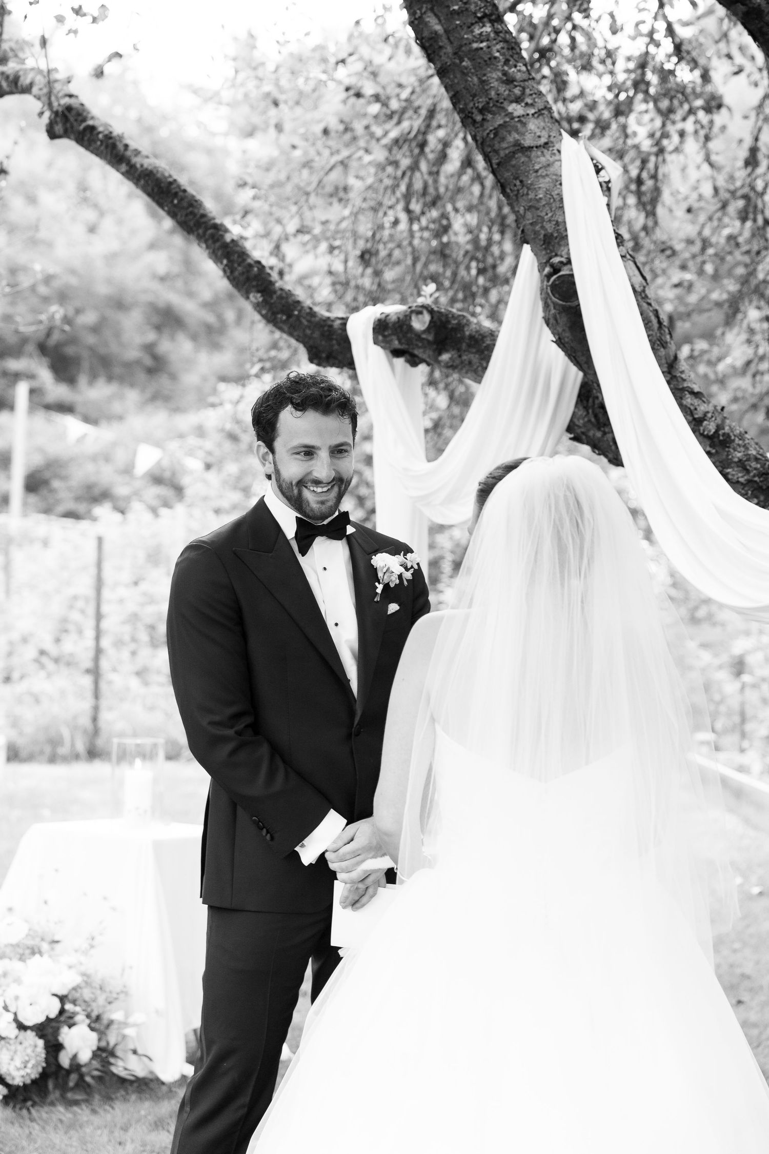 Black and white photo of groom smiling at bride during outdoor wedding ceremony beneath draped tree.