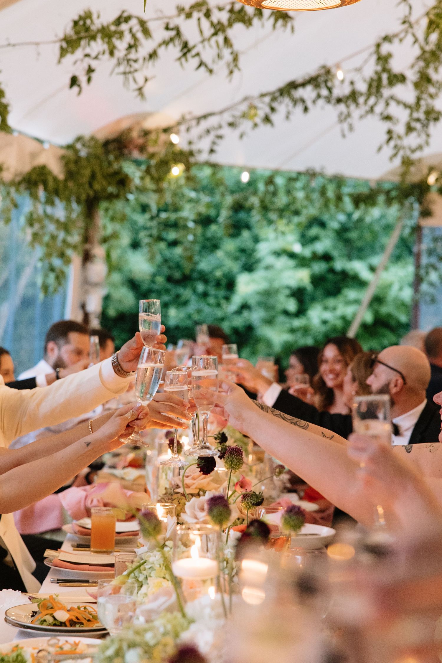Guests raising champagne glasses in a toast at an elegant outdoor wedding reception under a white tent with lush greenery.