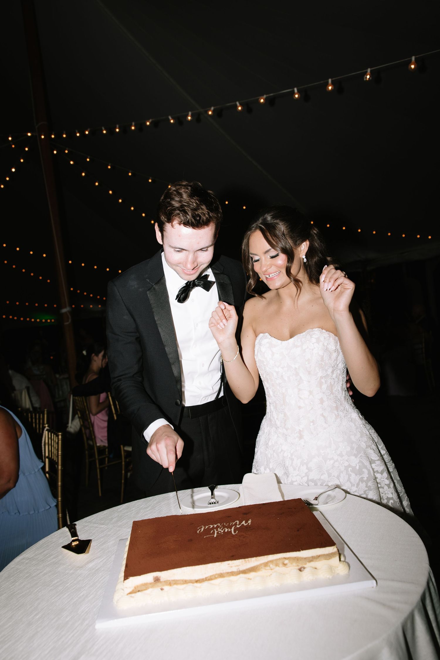 Bride and groom smiling while cutting their wedding cake under a tented reception with string lights.