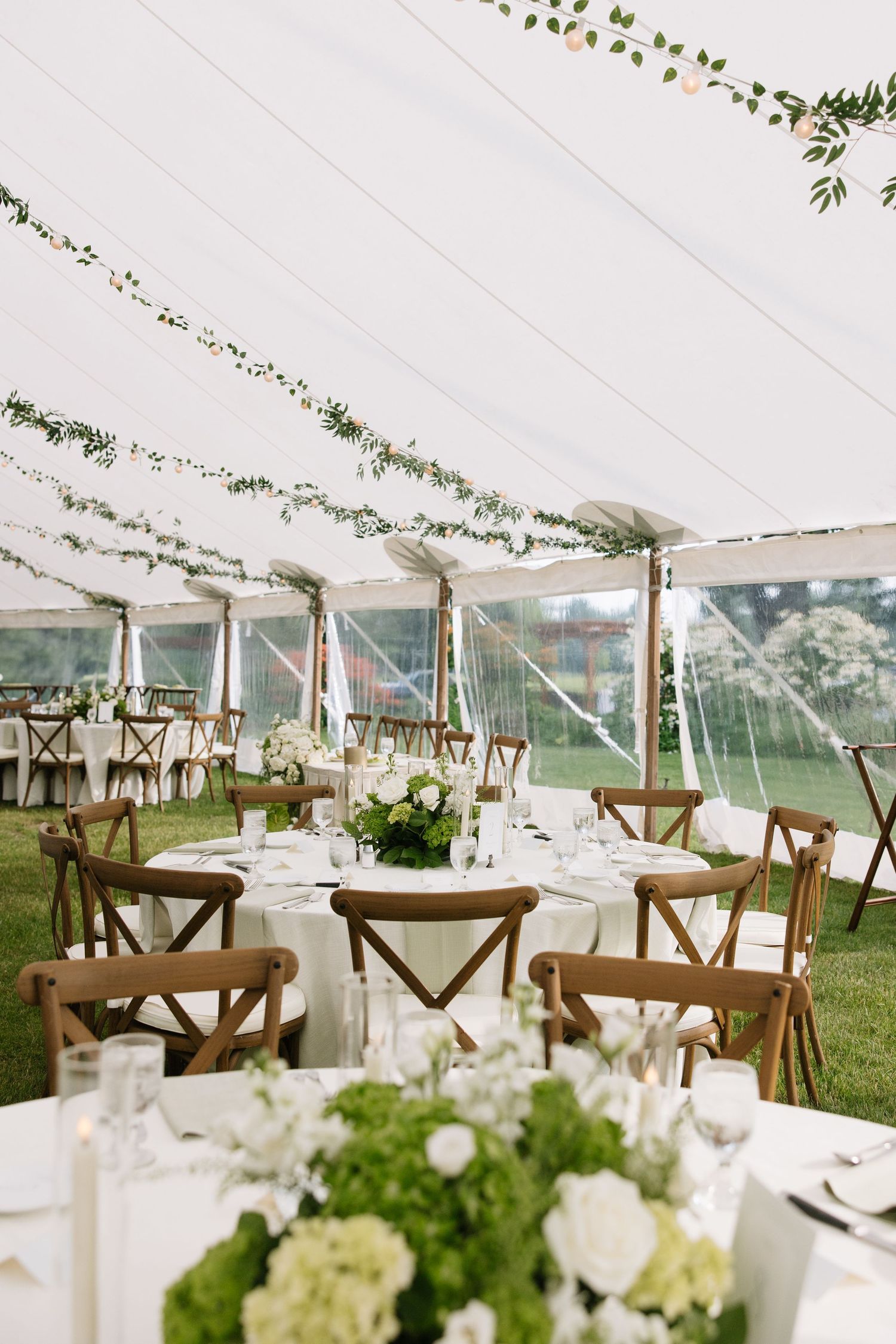 Elegant outdoor wedding reception tent with round tables, white floral centerpieces, wooden cross-back chairs, and greenery garlands.