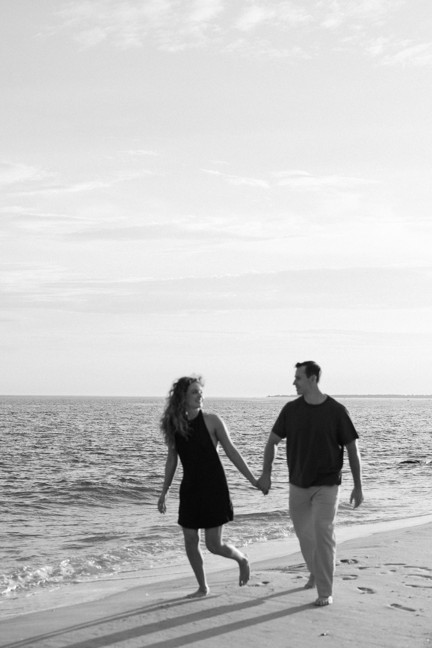 Black and white photo of a couple holding hands and walking along a sandy beach with ocean waves in the background.