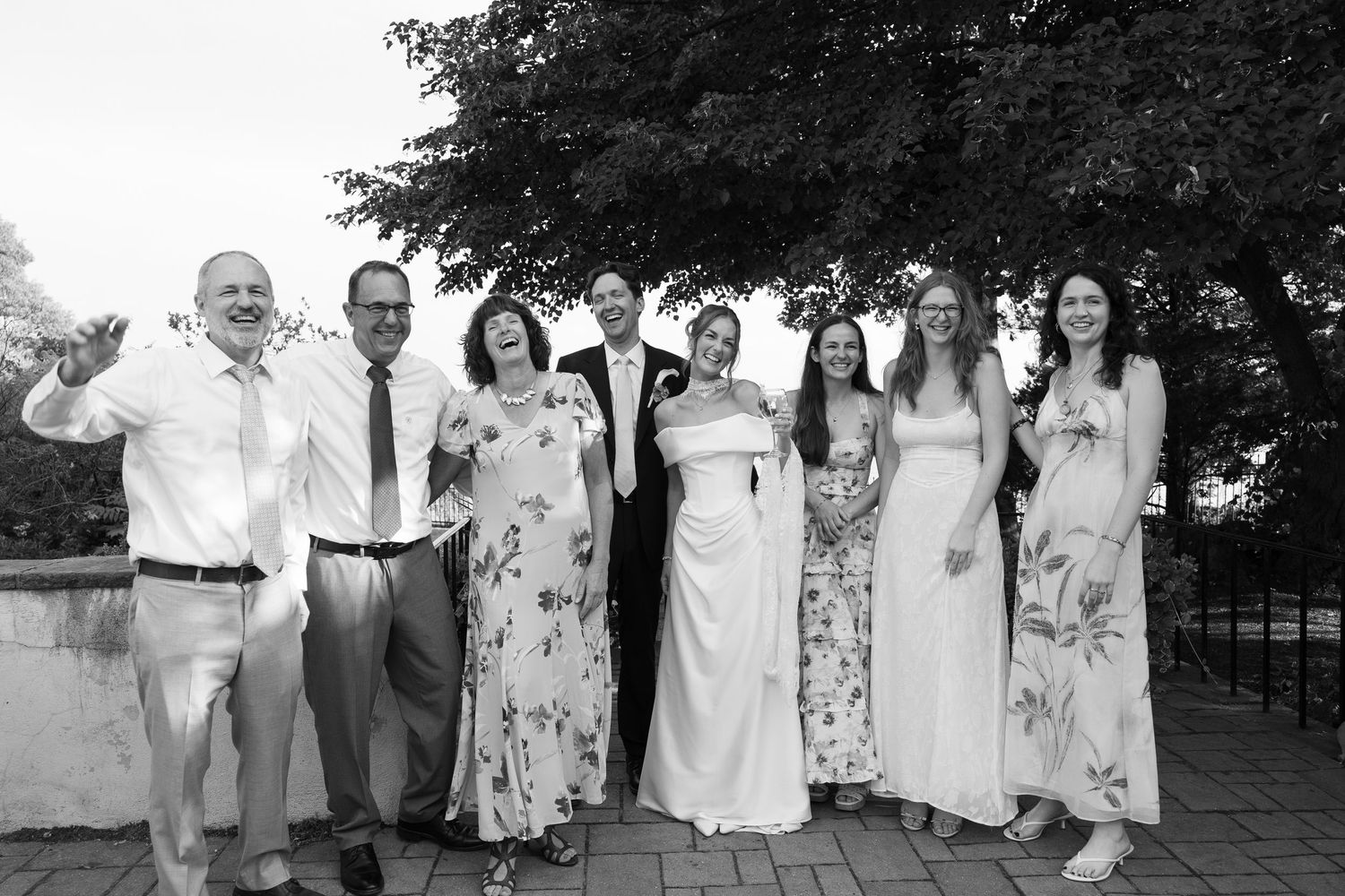 Black and white wedding party photo with bride, groom, and guests posing outdoors on brick patio near trees.