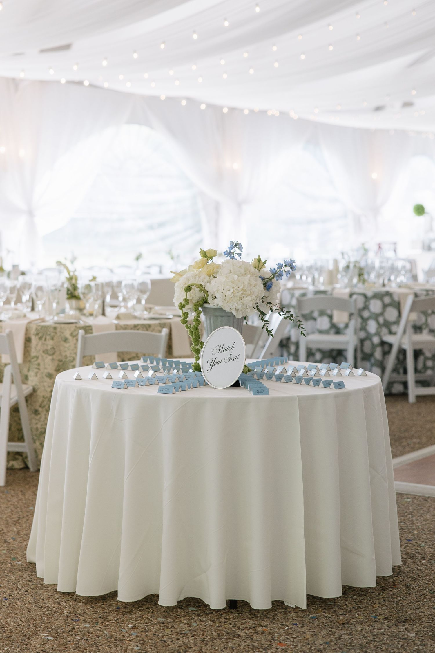 White tent wedding reception with round tables, floral centerpieces, and a escort card table with thank you sign.