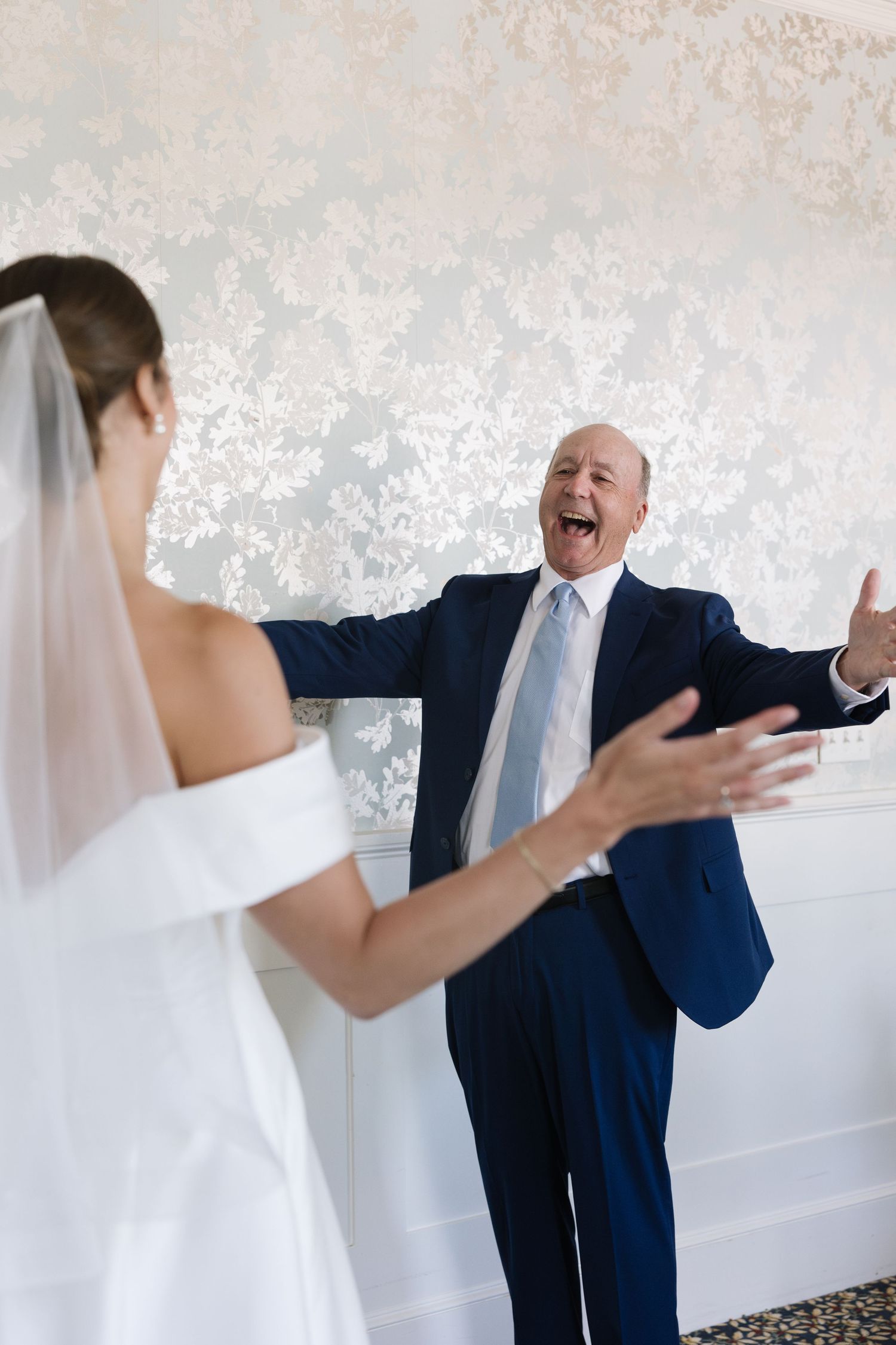Father reacts with joy and open arms seeing bride daughter in wedding dress during emotional first look moment.