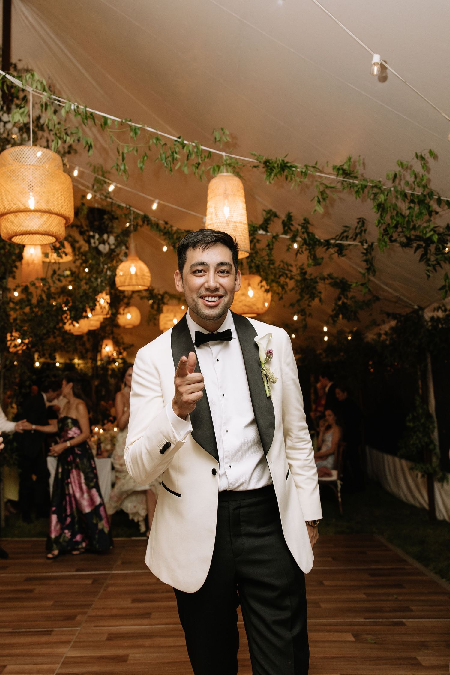 Smiling groom in white tuxedo jacket pointing at camera inside elegant tent reception with hanging lanterns and greenery.