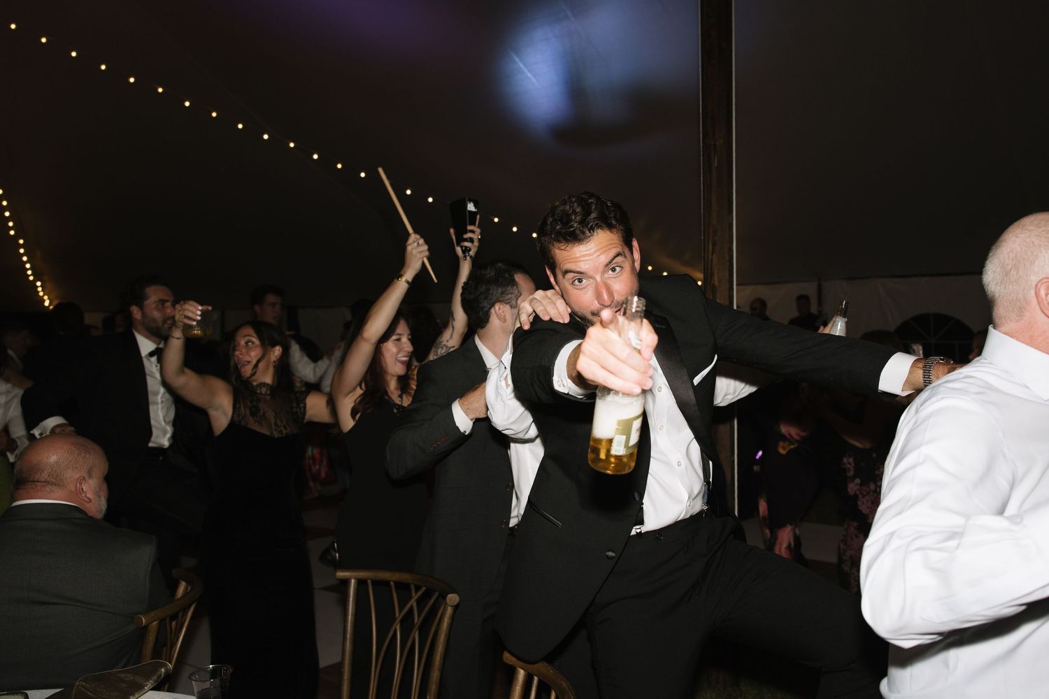 An energetic groomsman dances holding a beer bottle at a lively indoor wedding reception at night.