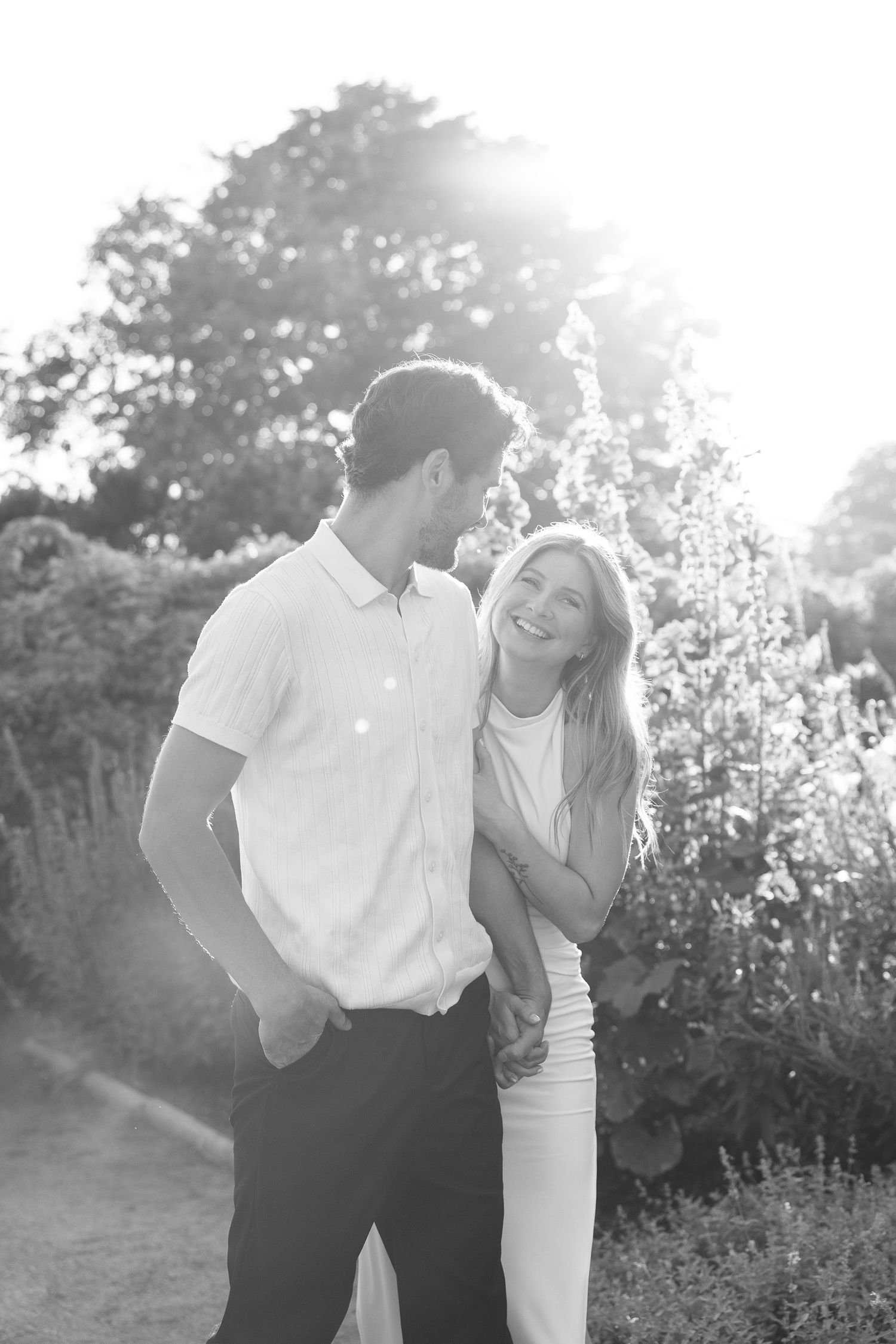 Black and white photo of a smiling couple holding hands in a sunlit garden with lush greenery.
