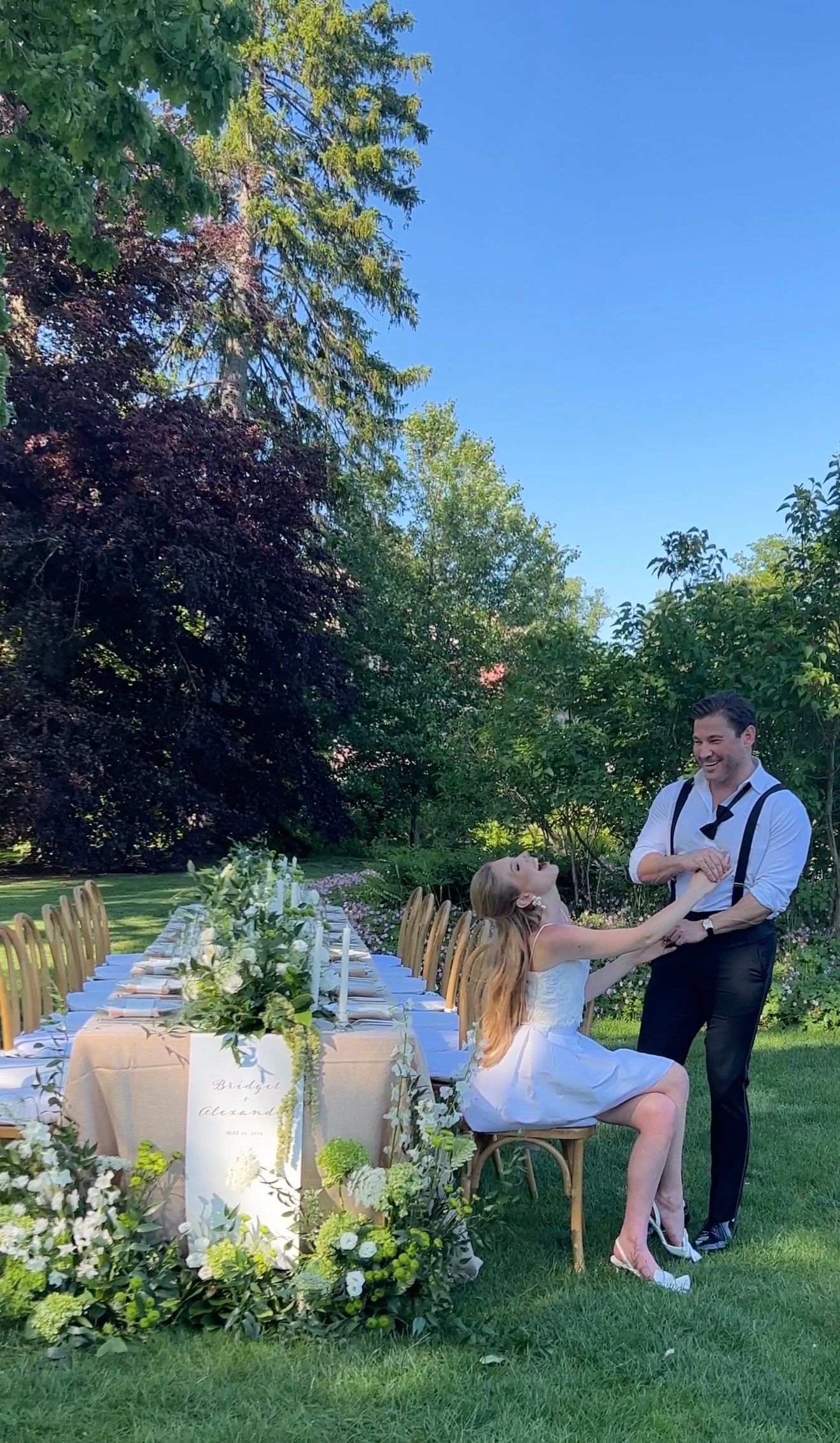 Couple laughing at elegant outdoor garden wedding reception with long floral table setting and gold chairs.