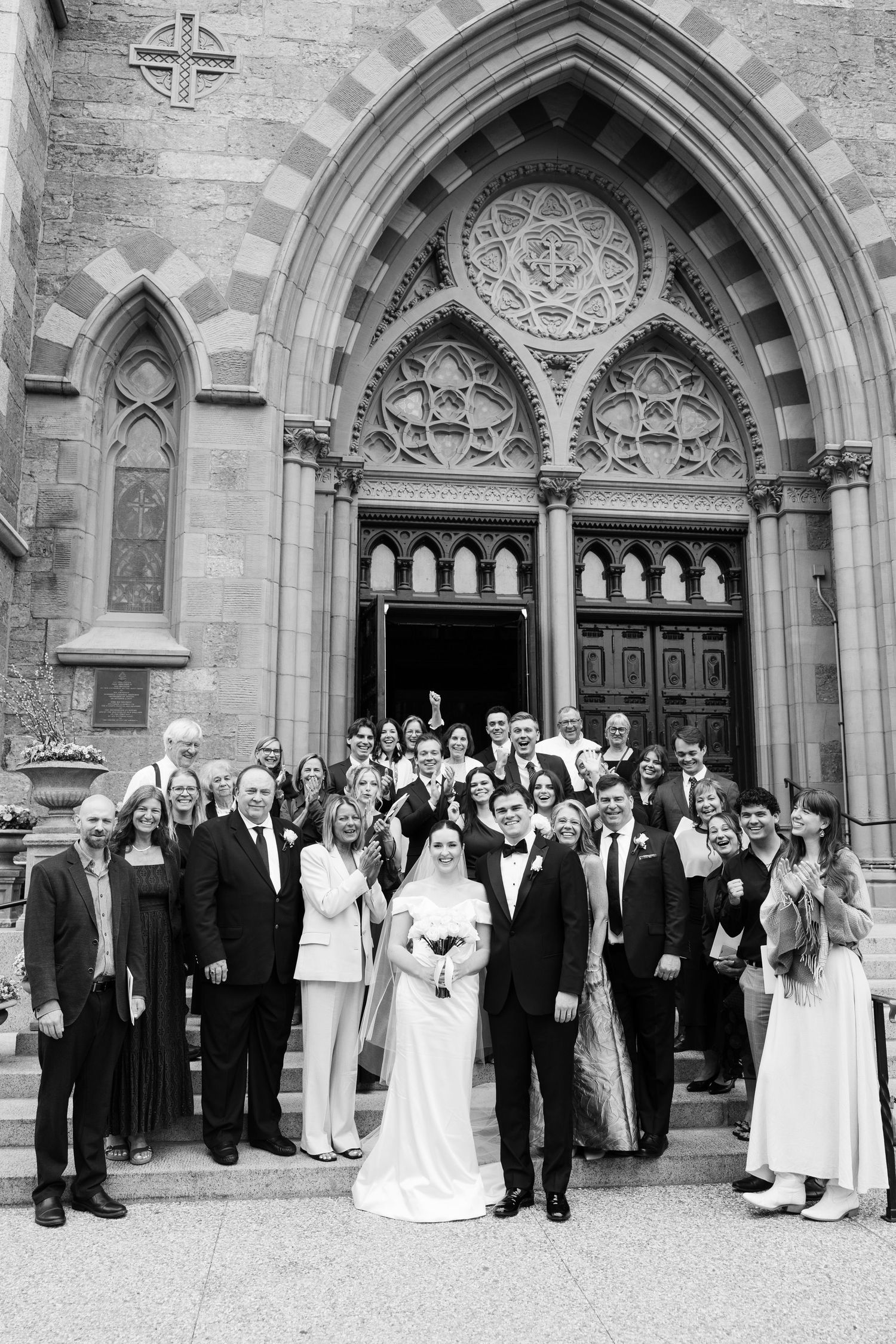 Wedding party poses on church steps in black and white photo outside gothic cathedral entrance.