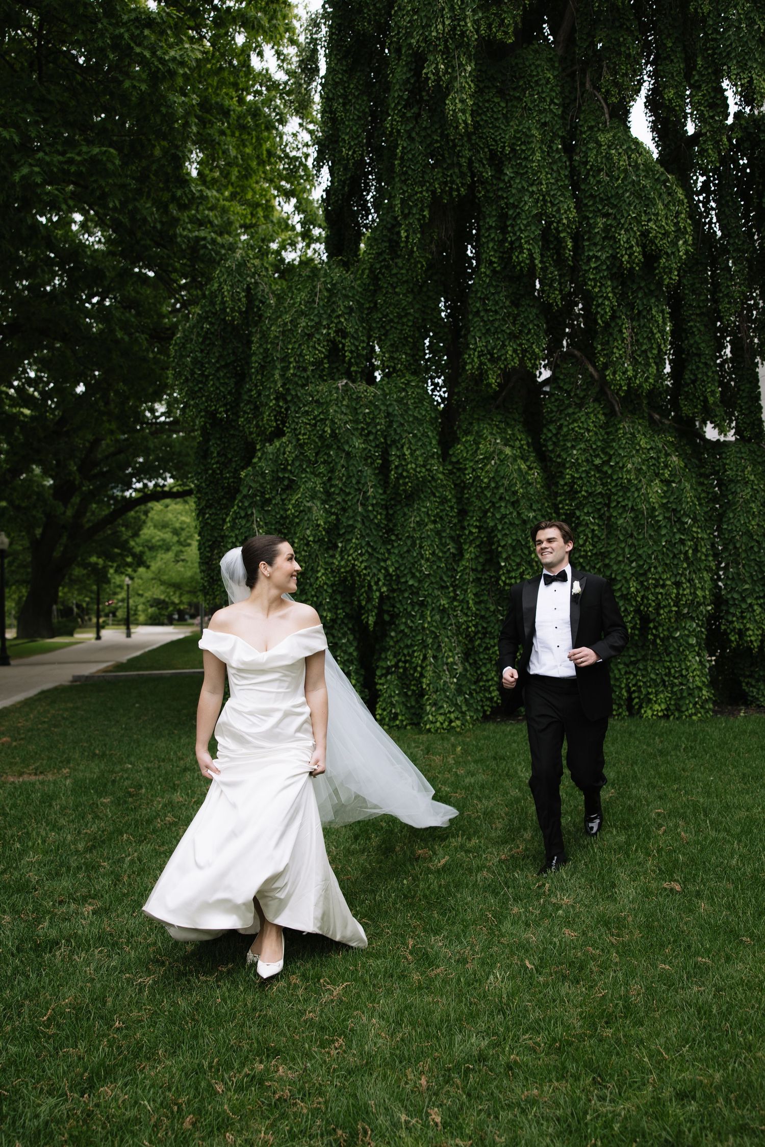 Bride in white off-shoulder gown and veiled groom in black tuxedo walk on lush green lawn near weeping tree.