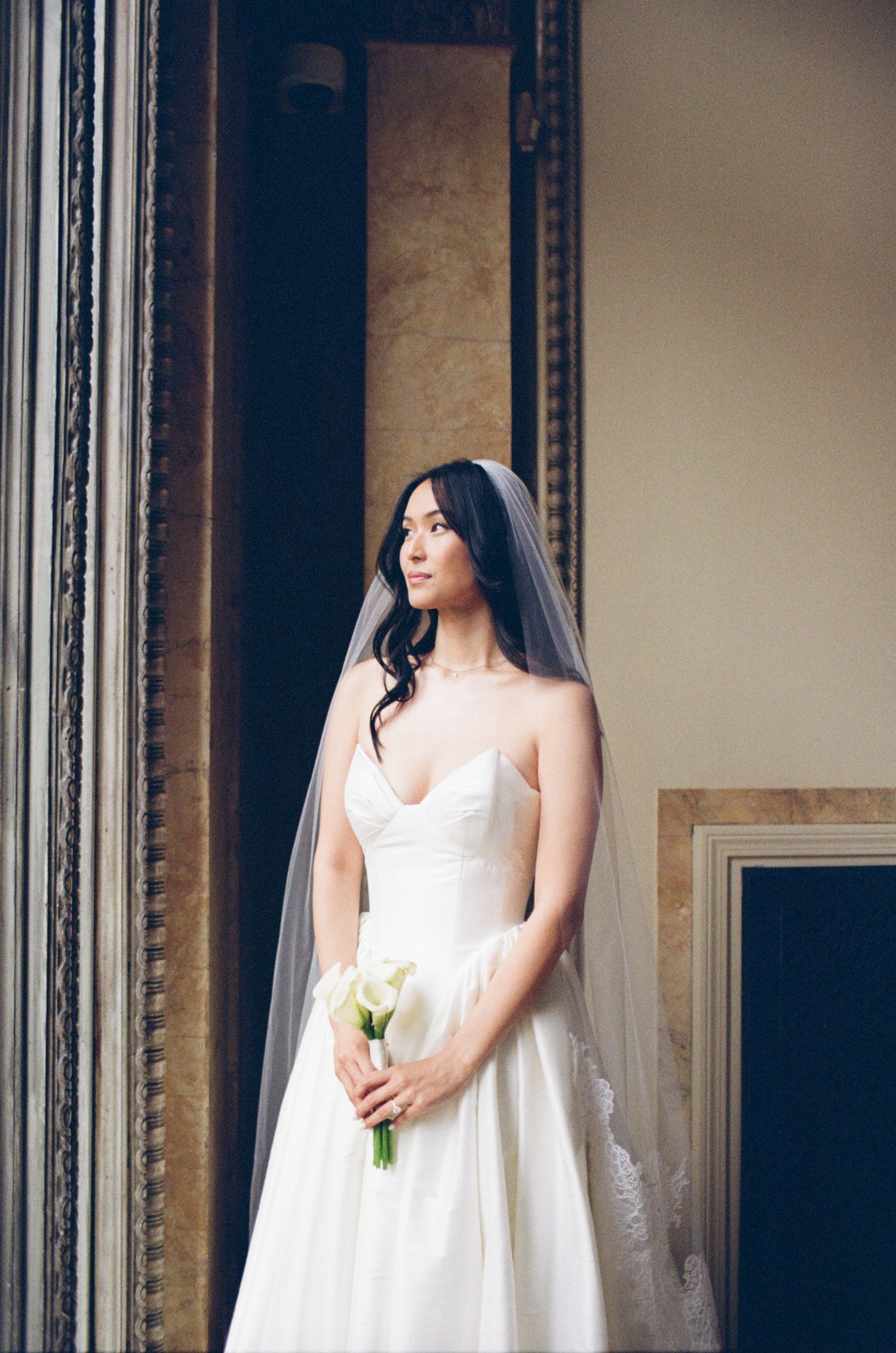 Elegant bride in strapless white gown and veil holding calla lily bouquet, standing in ornate venue doorway.
