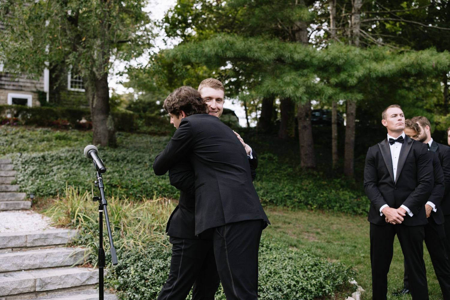 Wedding party members embrace in formal attire on stone steps surrounded by greenery.