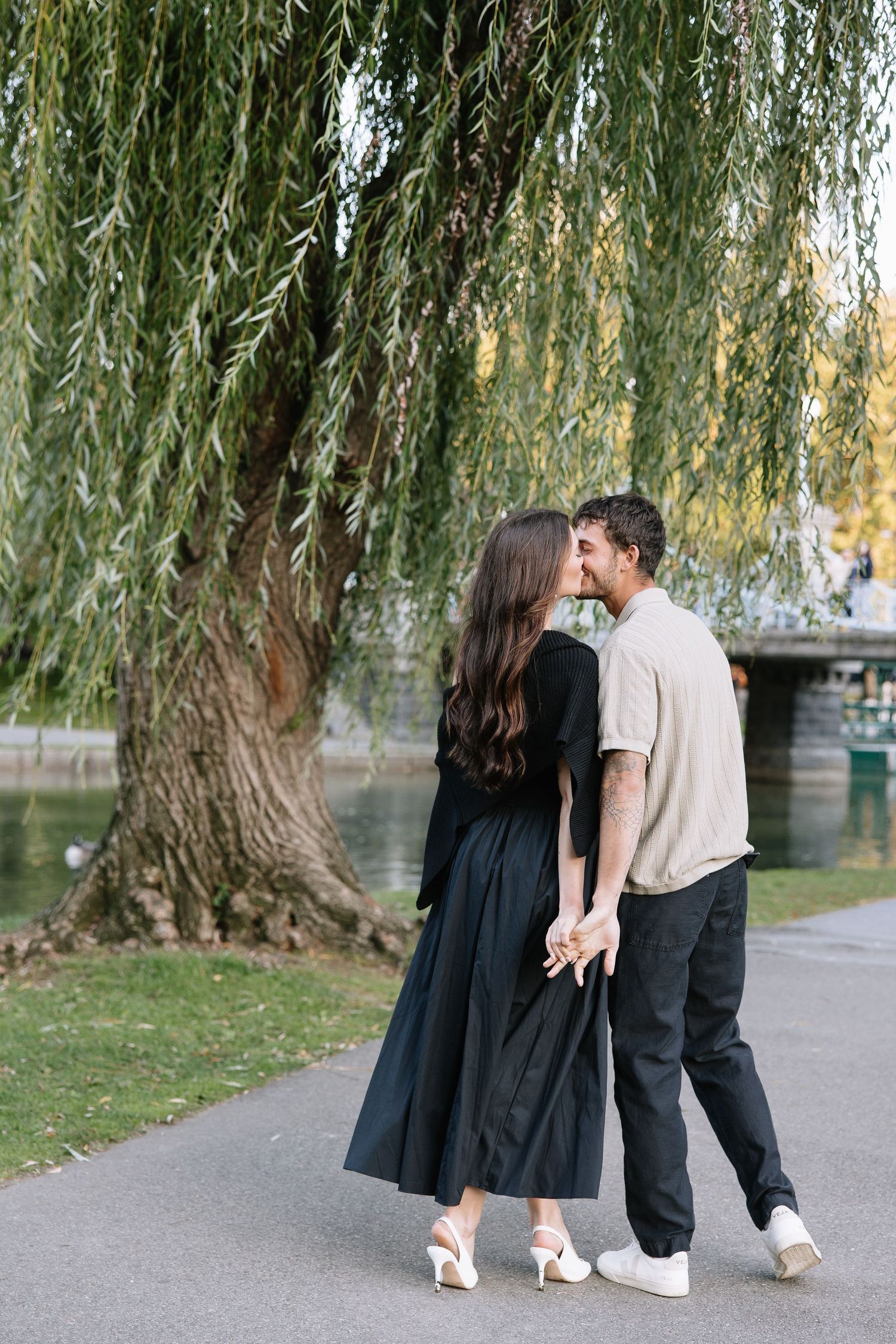 A couple walks beneath drooping willow branches along a park pathway at sunset.