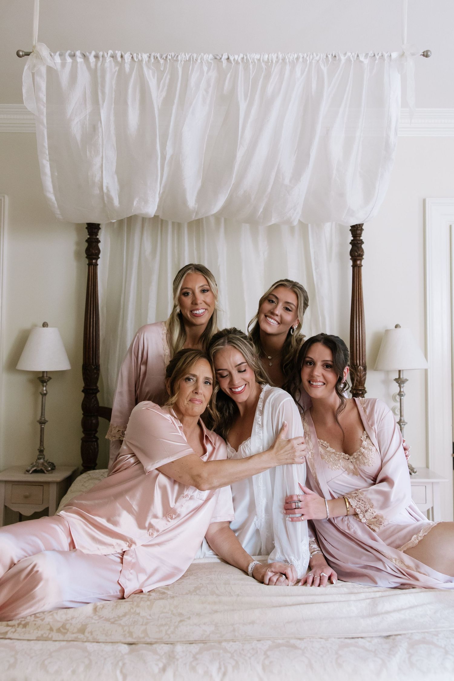 Bridesmaids wearing matching pink robes lounge together on a white bed during wedding preparations.