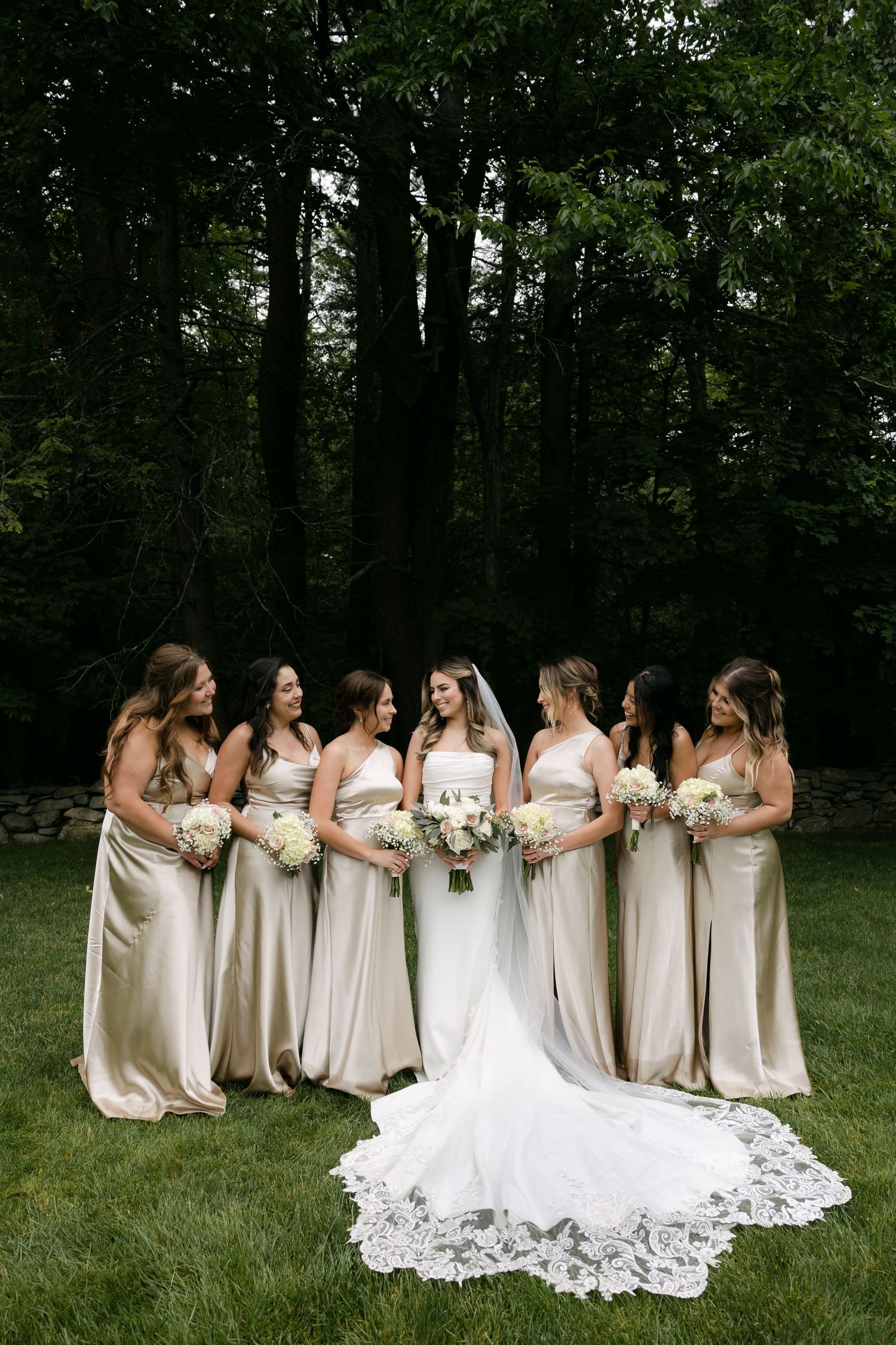 Bridesmaids in champagne dresses stand with bride in white gown on grassy lawn.