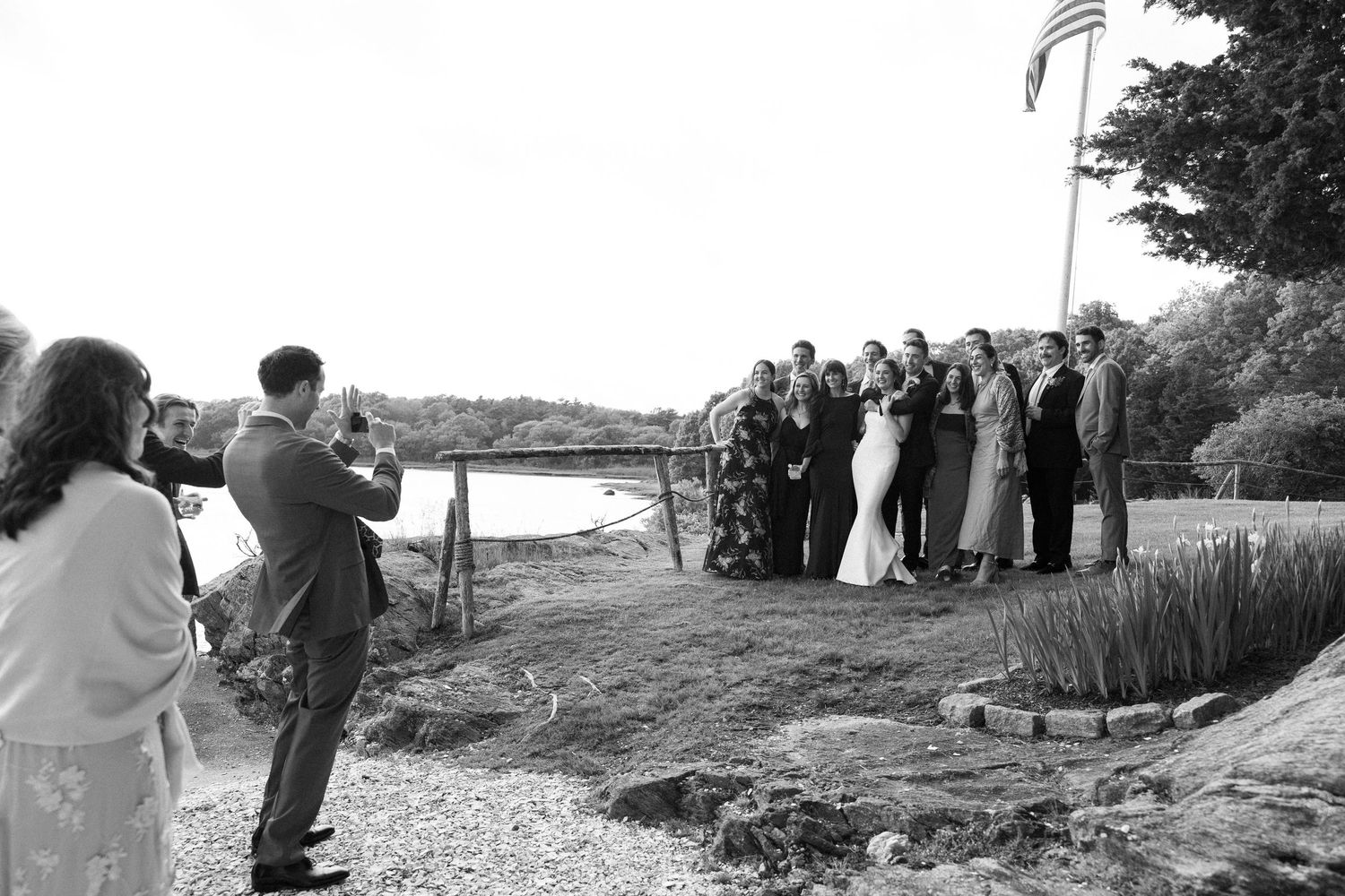 Black and white wedding photo of a photographer capturing a group portrait of the wedding party by a scenic waterfront.
