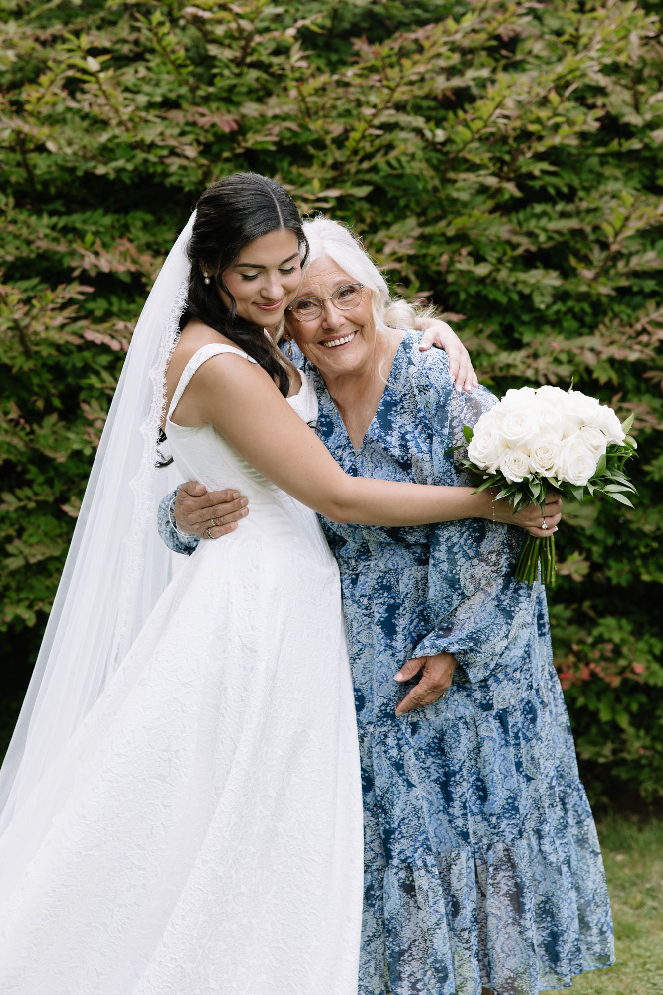 A bride in white gown and veil embraces an older woman in blue floral dress holding white bouquet outdoors.