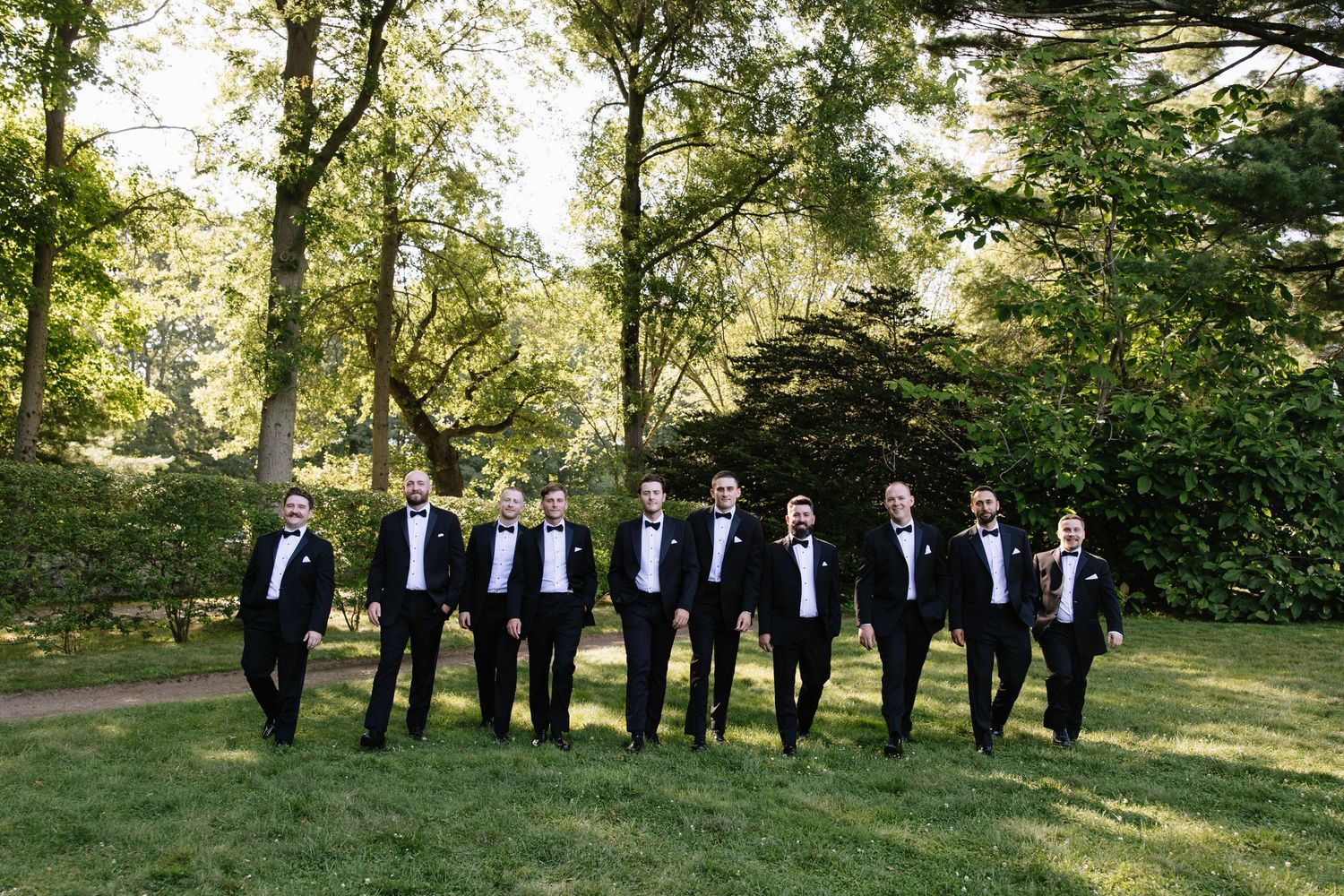 Groom and groomsmen in black tuxedos walking together on lush green lawn surrounded by trees.