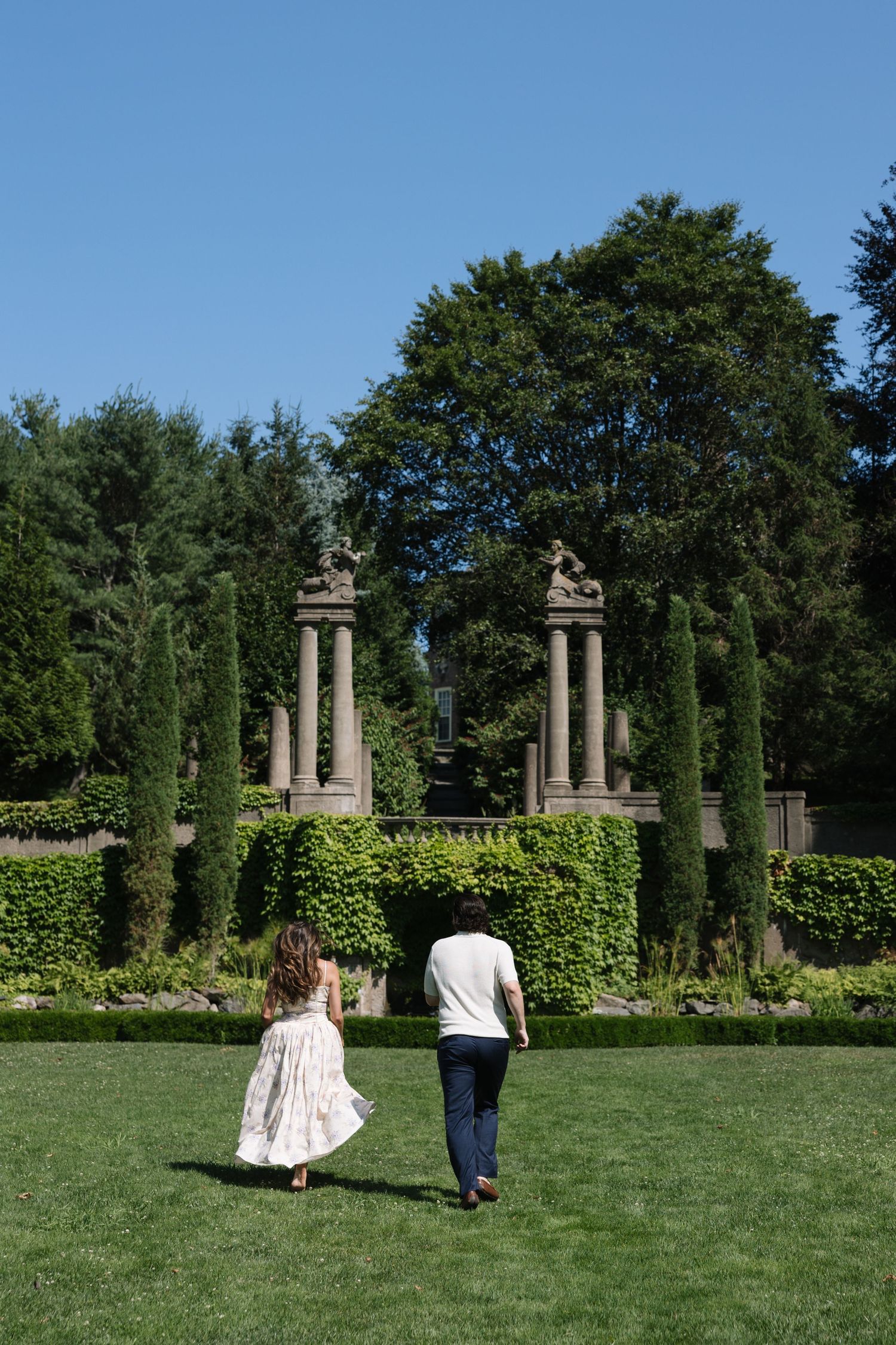 Couple walking hand in hand toward stone columns and lush green hedges in a formal garden on a sunny day.