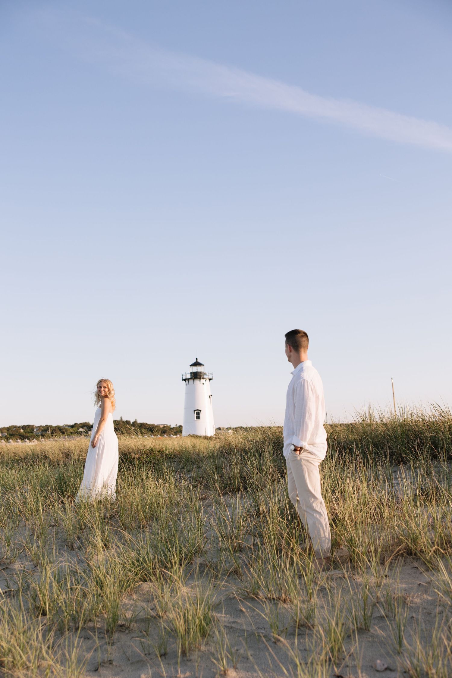 Couple in white attire standing on sandy dunes near a lighthouse under a clear blue sky.