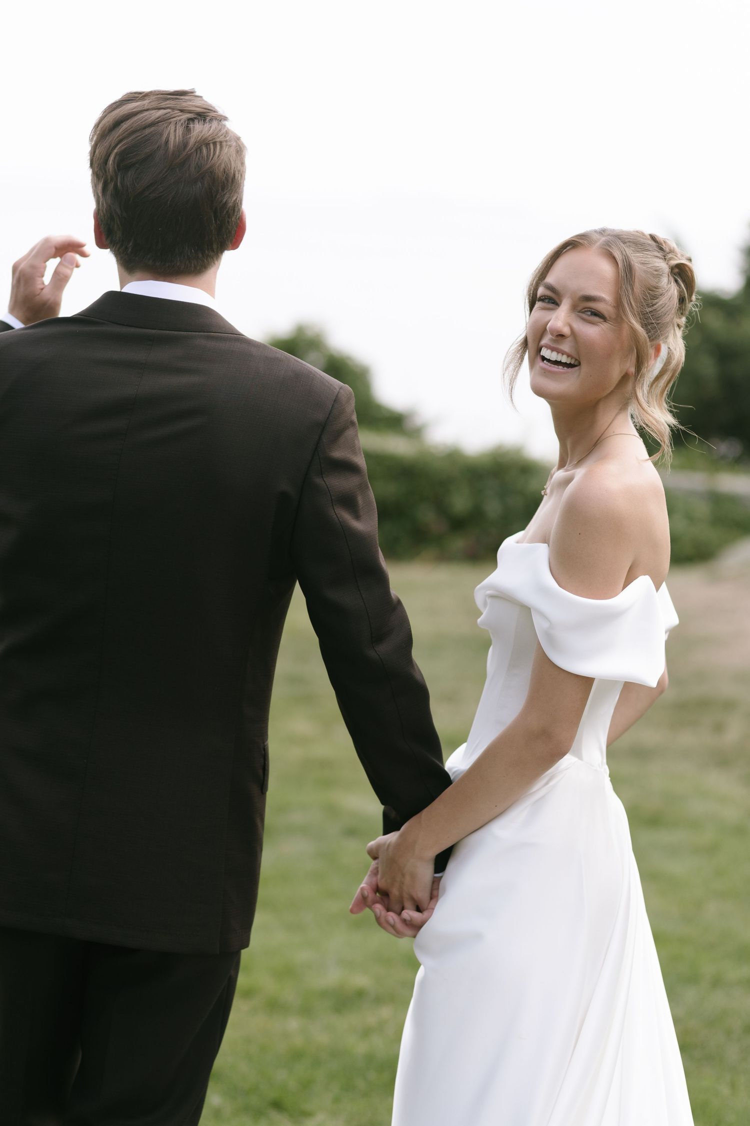 Smiling bride in an off-the-shoulder white wedding gown holds hands with her groom in a dark suit on a grassy lawn.