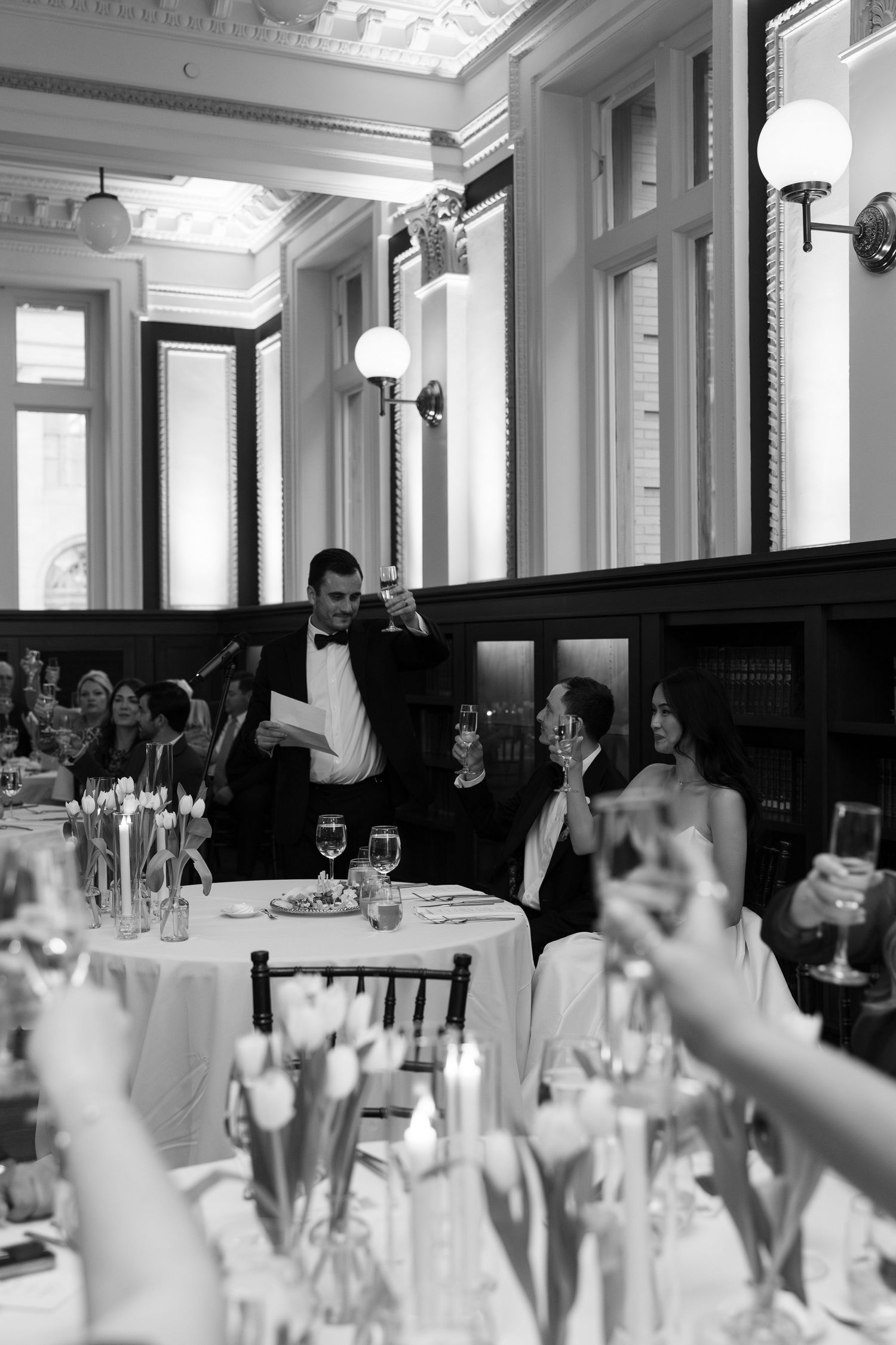 Black and white wedding reception toast with groom raising champagne glass while guests celebrate in elegant ballroom.