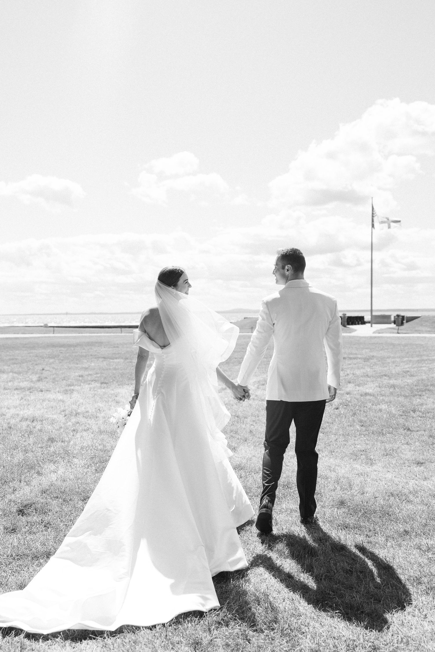 Black and white photo of bride and groom holding hands walking toward the ocean on their wedding day.