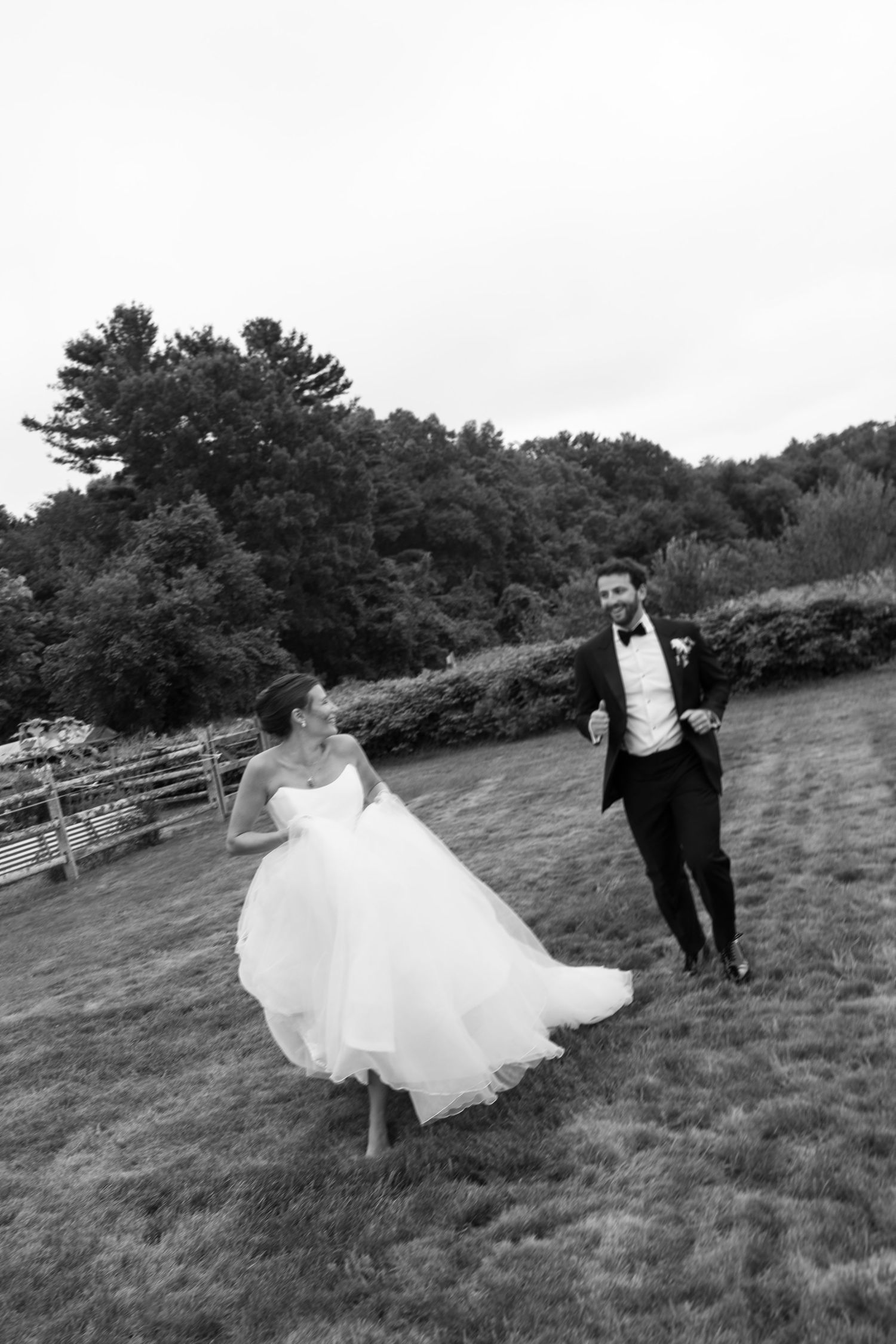 Black and white photo of bride and groom running joyfully across a grassy field after wedding.