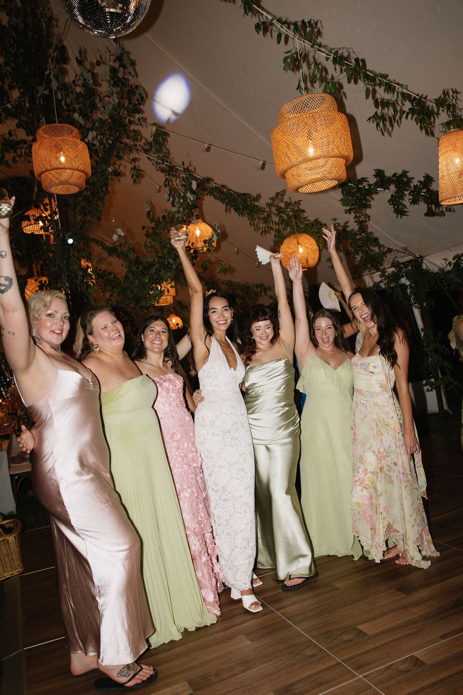 Bride and bridesmaids in mismatched pastel gowns celebrate joyfully under rattan lanterns at a garden wedding reception.