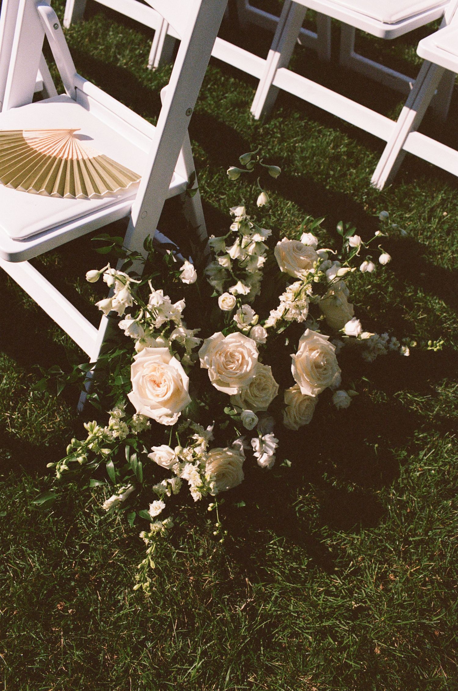 White roses and delicate blooms arranged on green grass beside white folding chairs at an outdoor wedding ceremony.