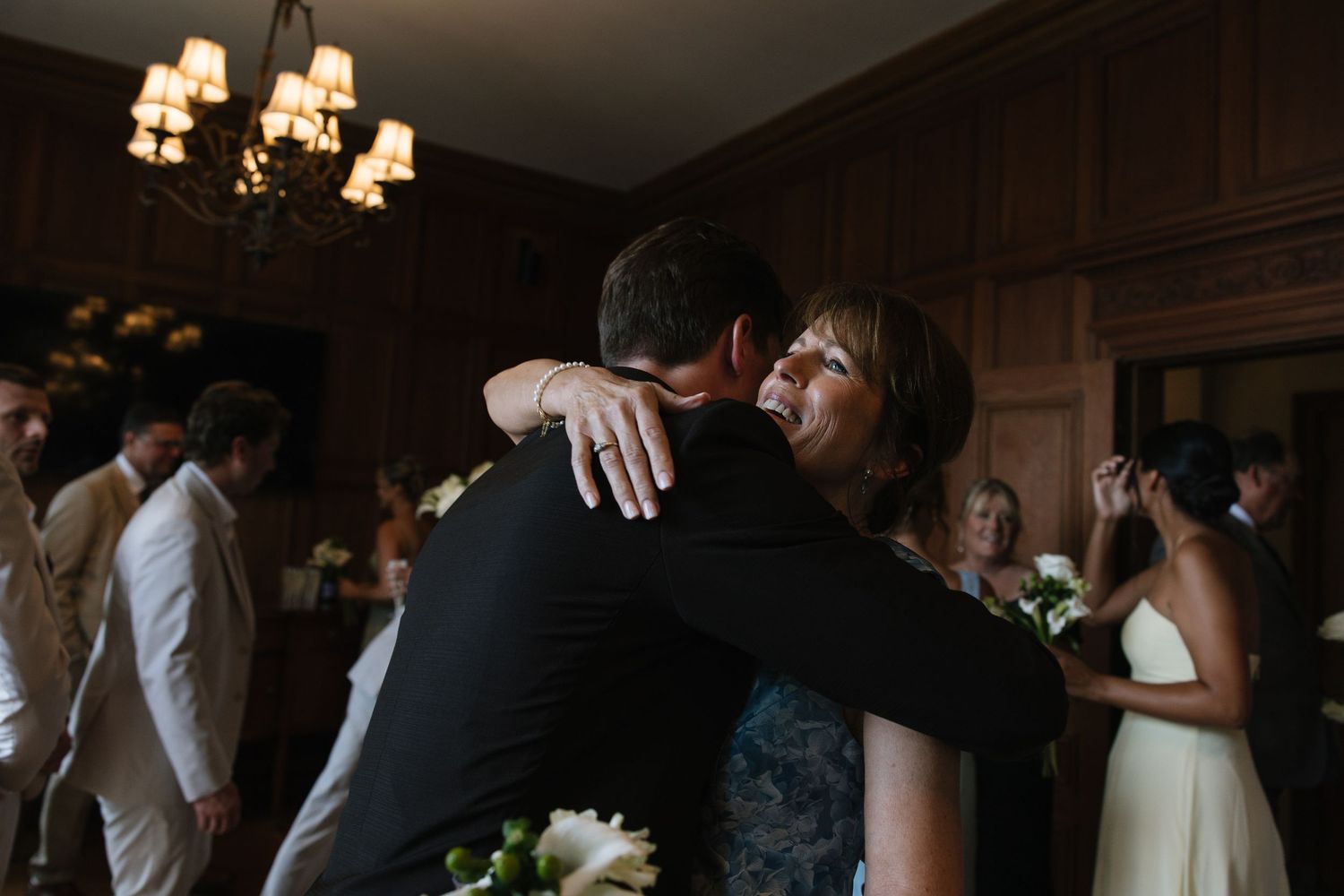 A woman hugs a groom in a dark suit at a wedding reception in an elegant wood-paneled room.