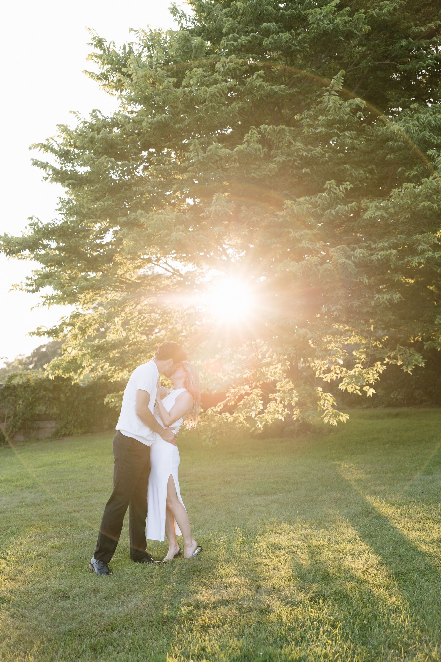 Couple embracing in golden sunset light in a lush green park, woman in white dress and man in black pants.