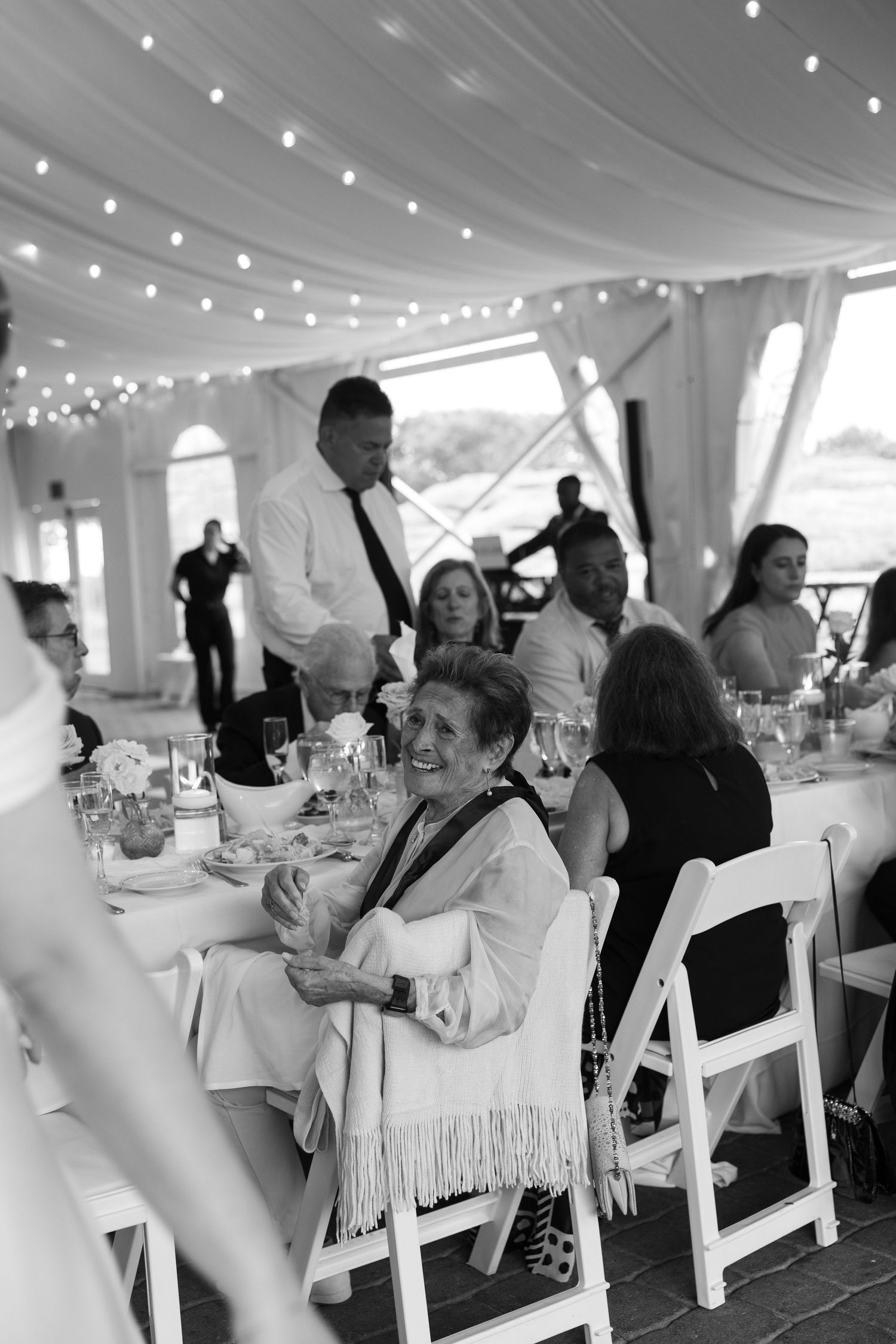 Black and white photo of wedding reception guests enjoying dinner under string lights.