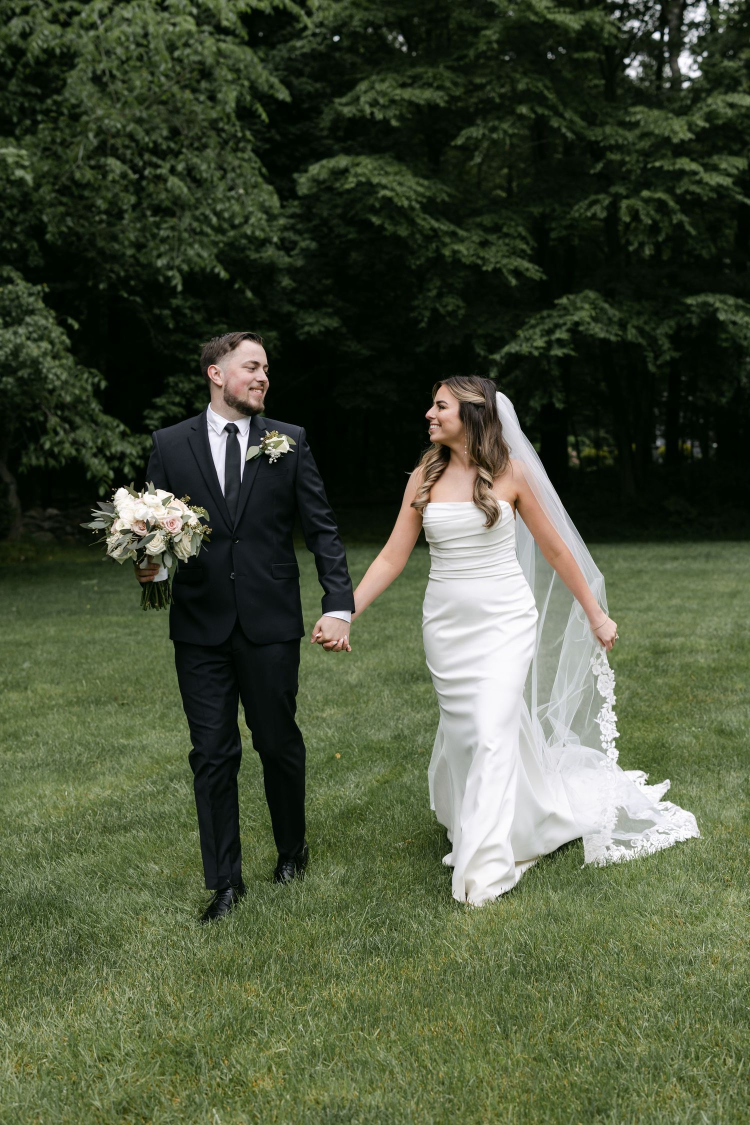 Newlyweds walk hand in hand across sunny green lawn.