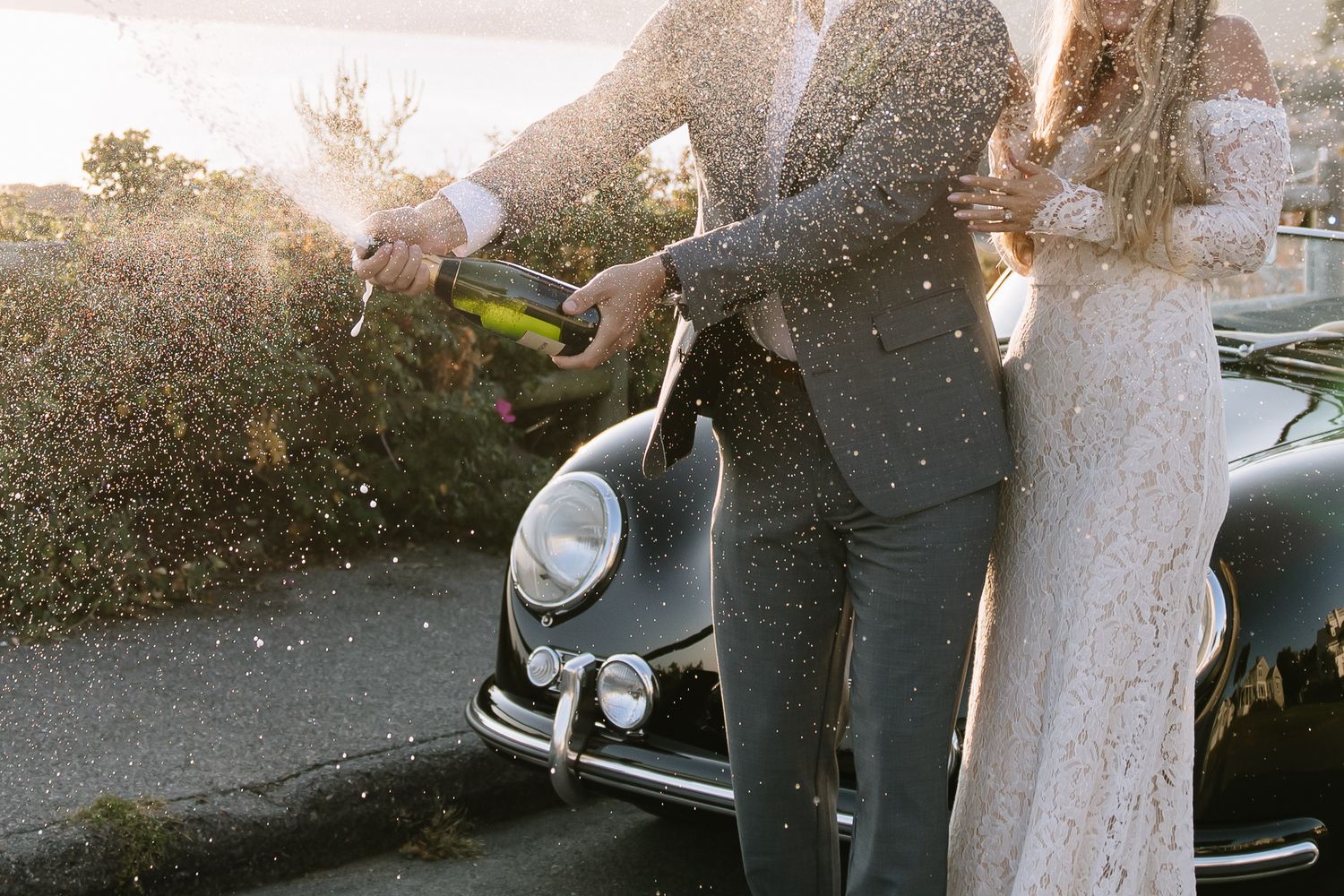 A couple stands next to their vintage black convertible car in evening light.