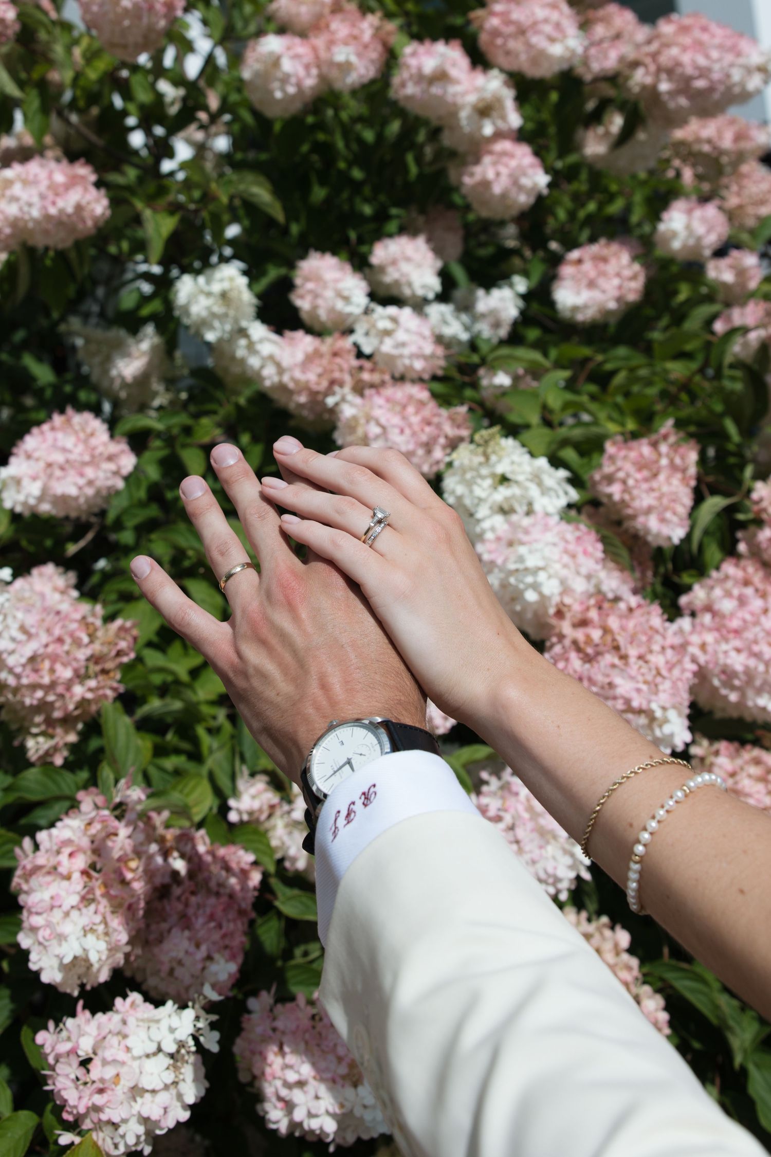 Two hands with wedding rings rest together among pink and white hydrangea flowers, one wearing a white watch.