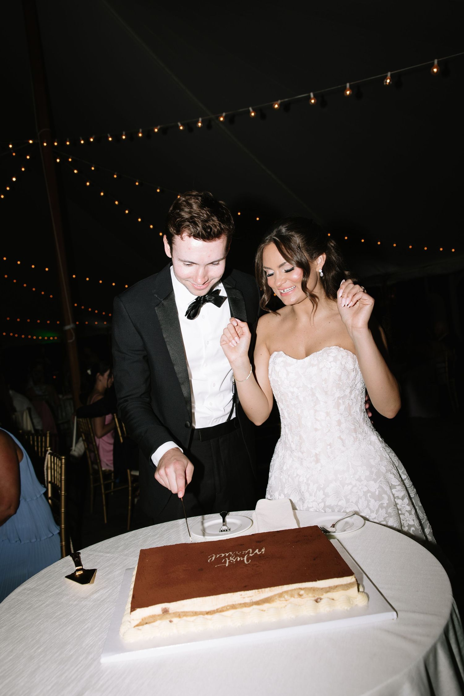 Bride and groom smiling while cutting their wedding cake under a tented reception with string lights.